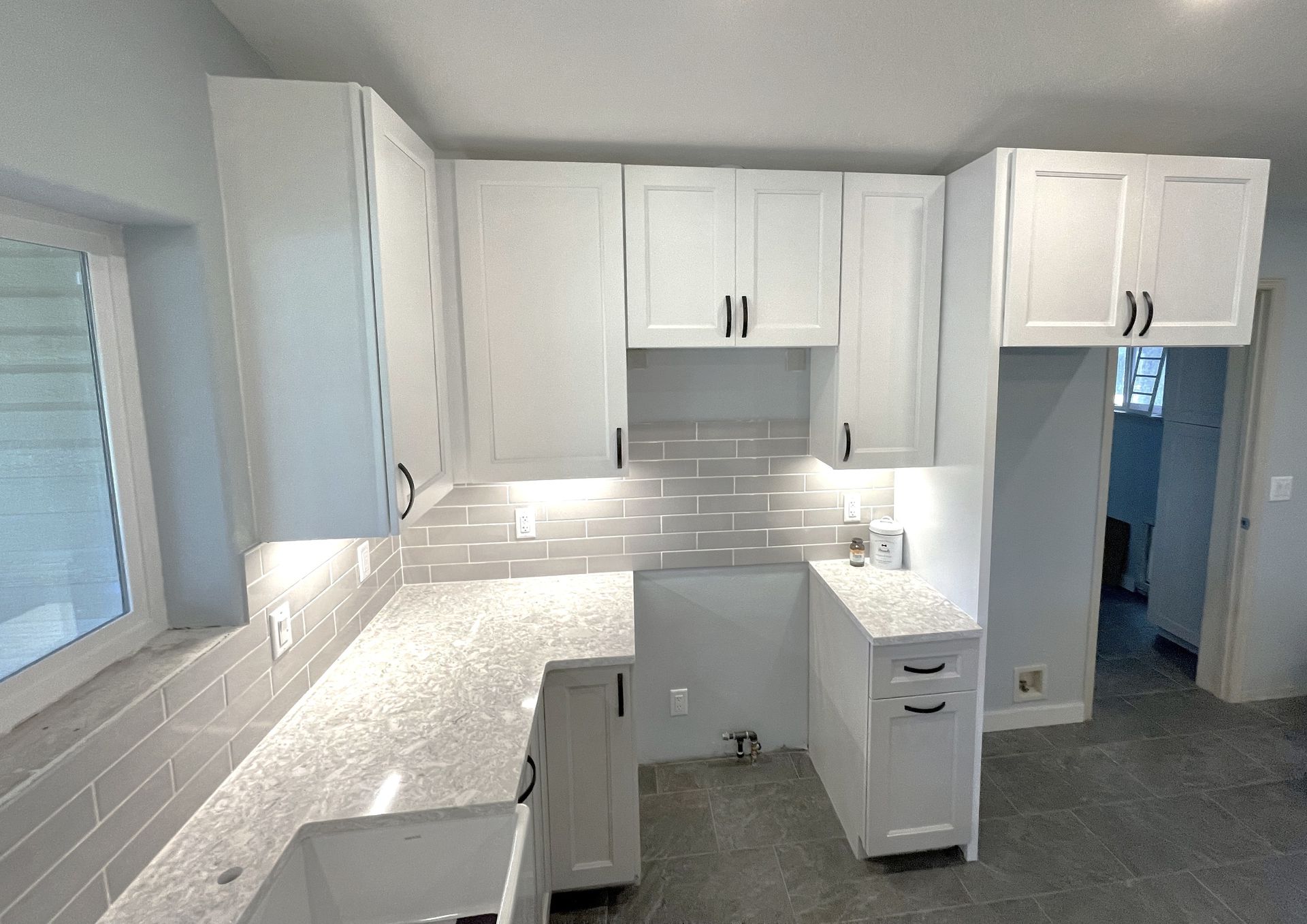 White kitchen cabinets with gray countertops and backsplash, with a window and a doorway.