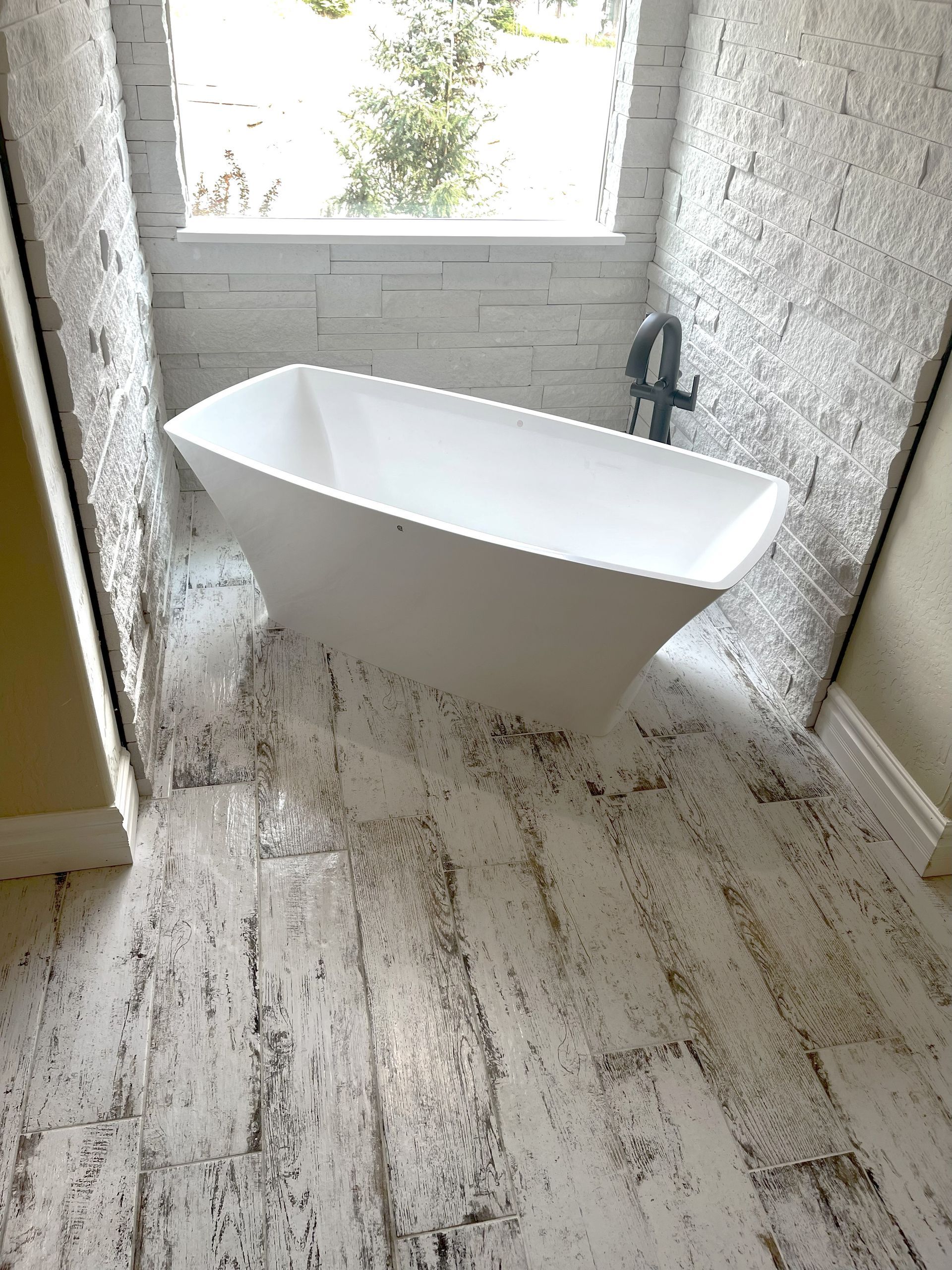 White freestanding bathtub in a bathroom with textured white stone walls and distressed white wood-look floor.