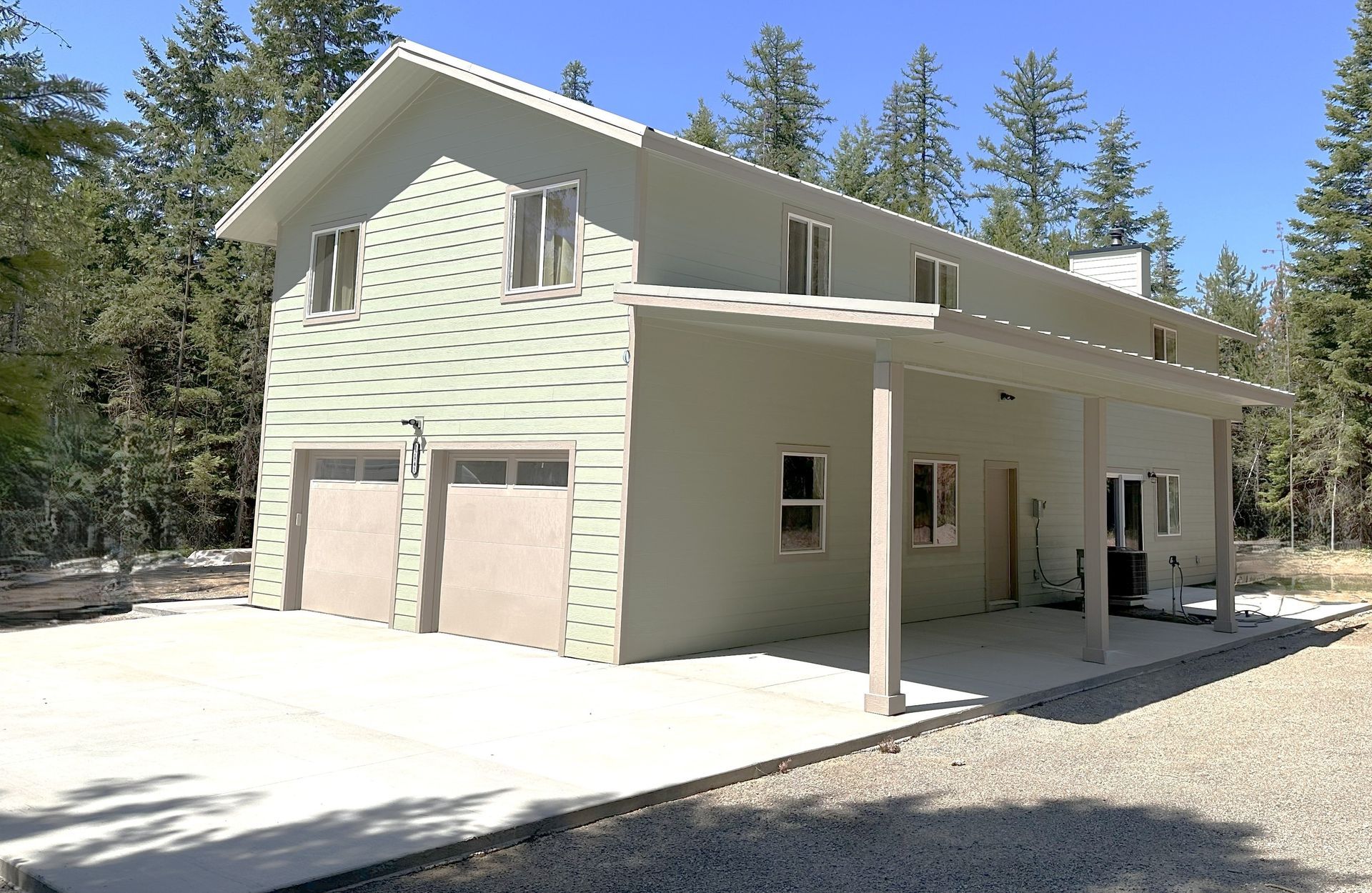 Two-story light green building with two garage doors and carport; gravel driveway, set in a forest.