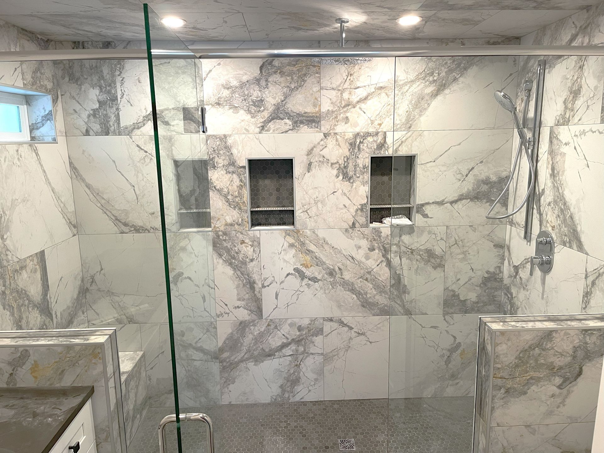 A mostly white marble-tiled shower with glass enclosure, built-in shelving, and chrome fixtures.