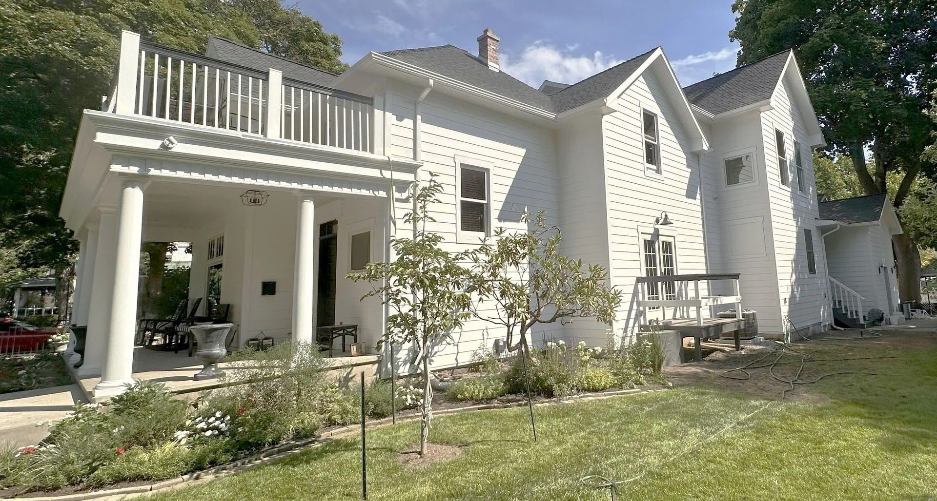 White two-story house with pillars and balcony. Green lawn, trees, and garden in front.