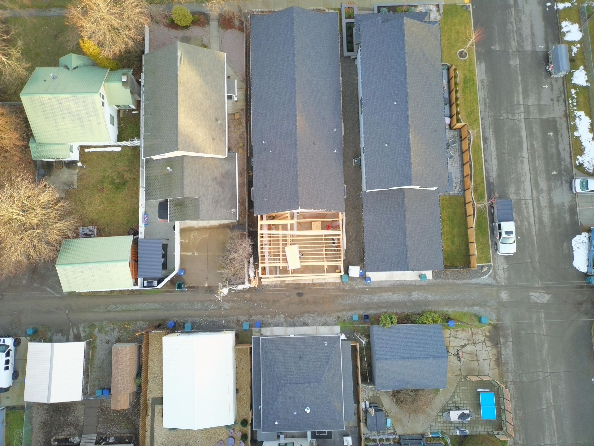 Overhead view of several houses with dark roofs. One has exposed wooden framing, suggesting construction.