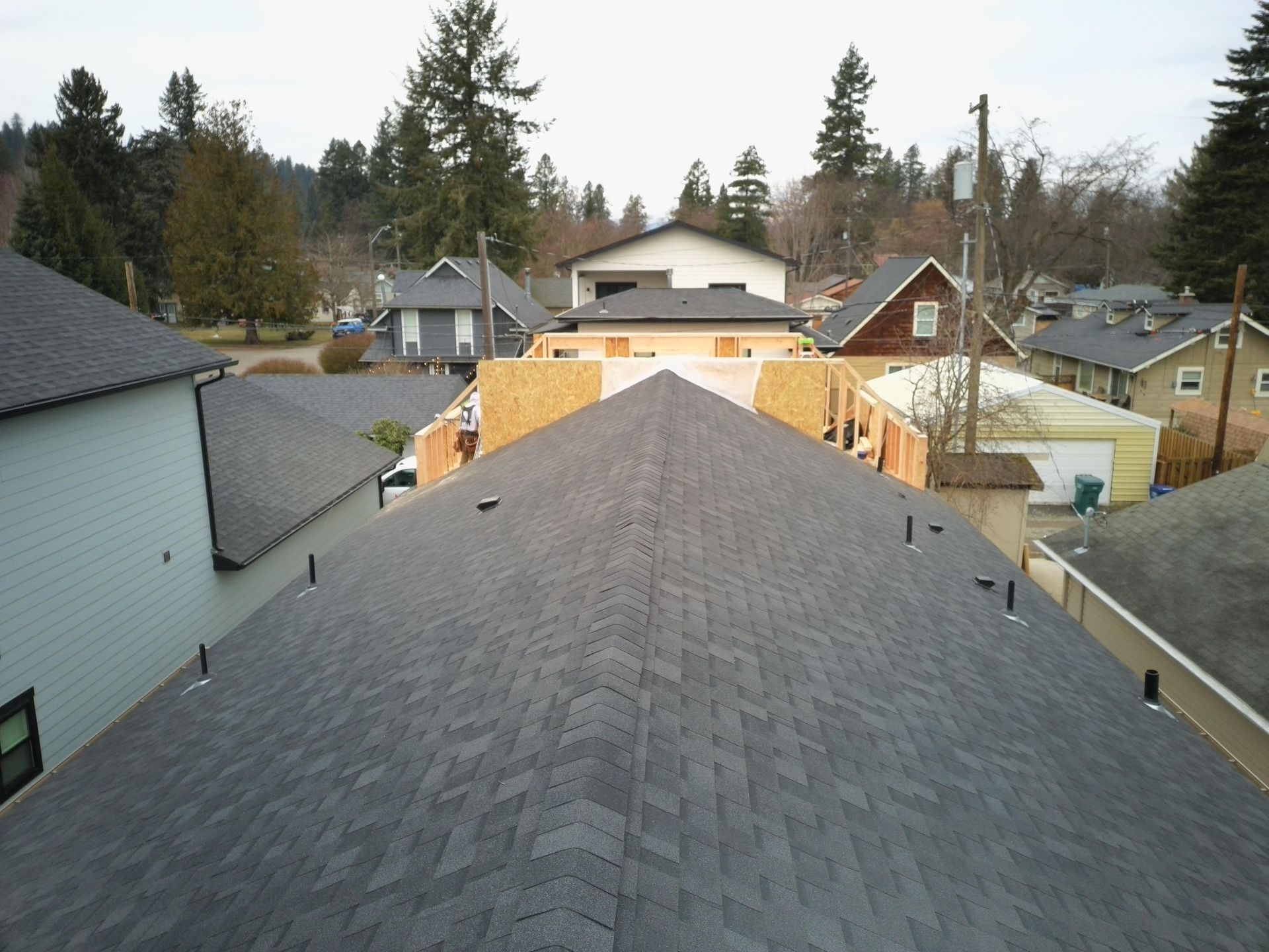 Roofs of houses, one with new construction, in a residential area. Gray asphalt shingles dominate the view.