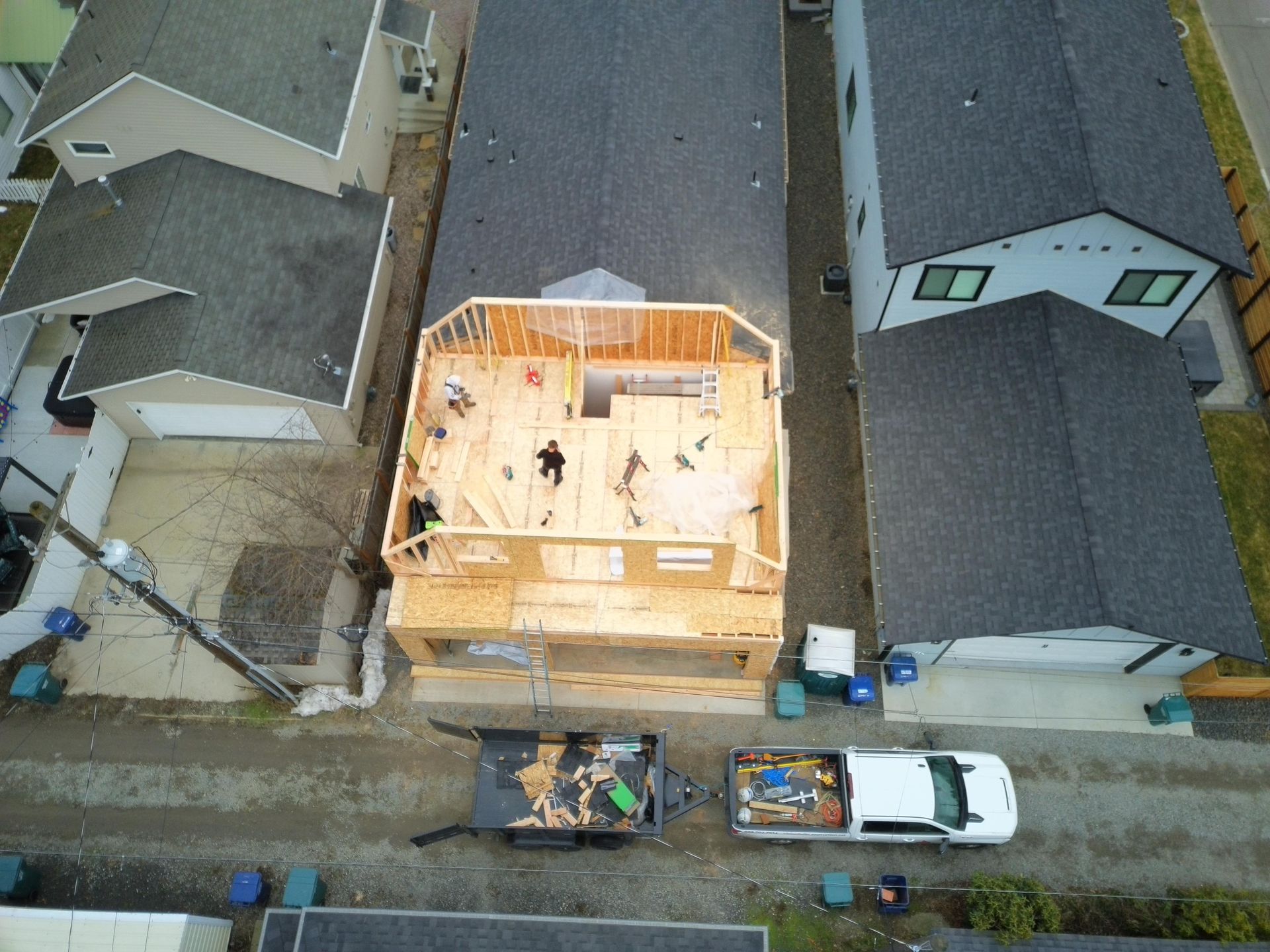 Aerial view: House under construction, workers on framing. Trailer and truck parked on gravel.