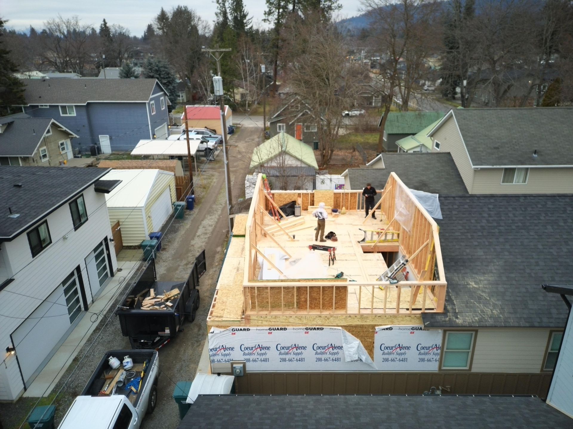 Construction of a second story addition on a house, workers on the wooden frame, with surrounding houses and a truck.