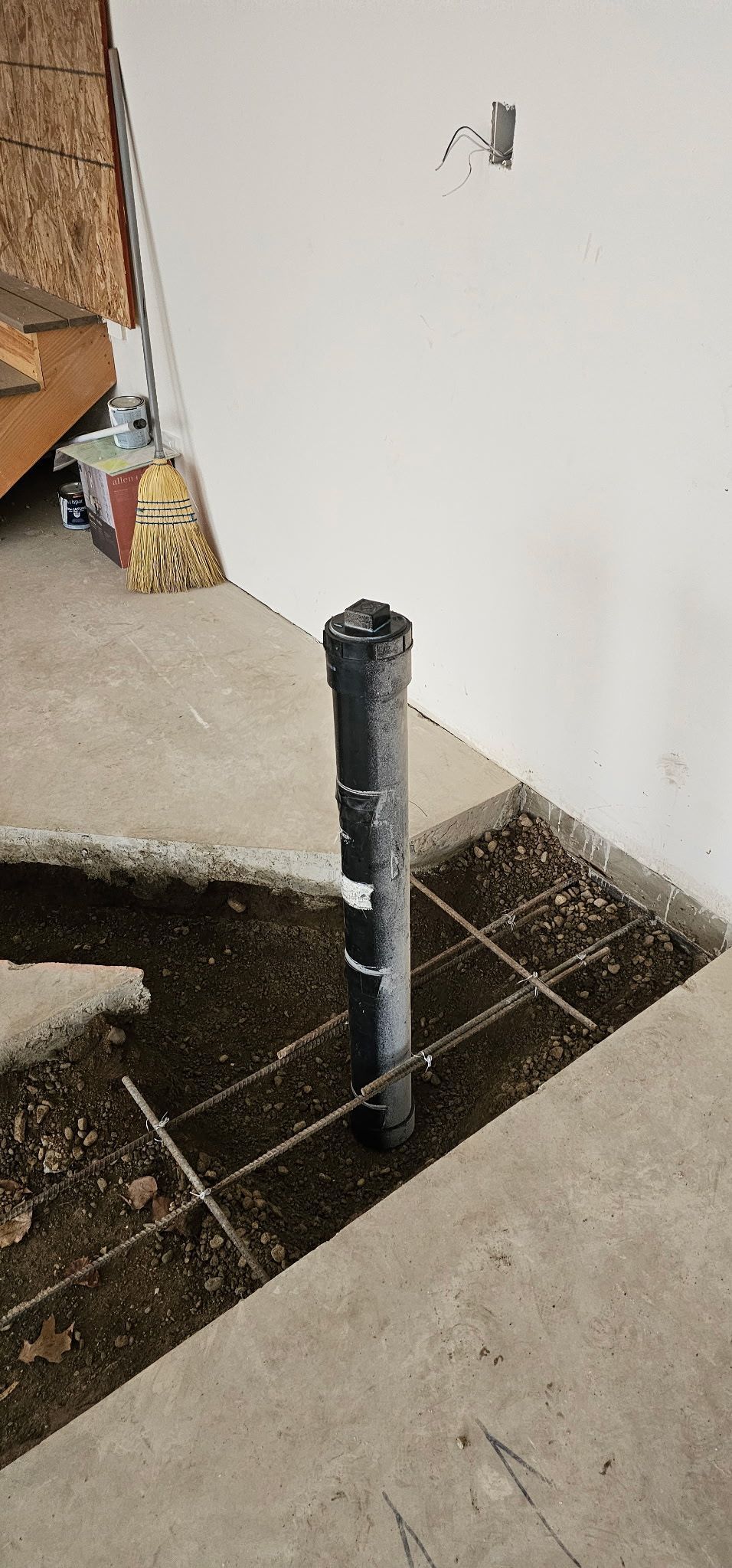 A black pipe stands in a square cut in a concrete floor, next to a white wall.
