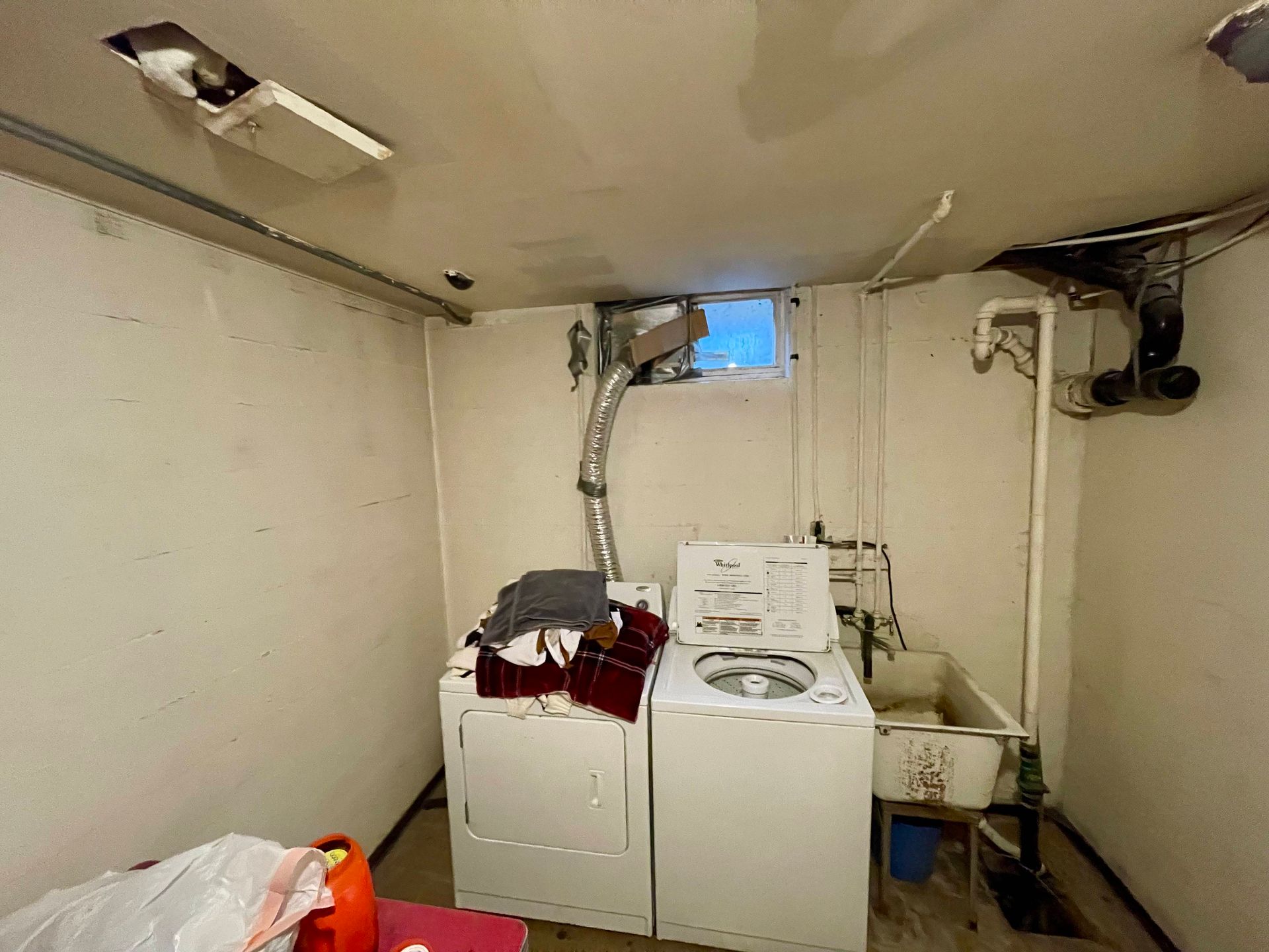 Laundry room with washer, dryer, sink, and small window. White walls, exposed pipes, and damaged ceiling.