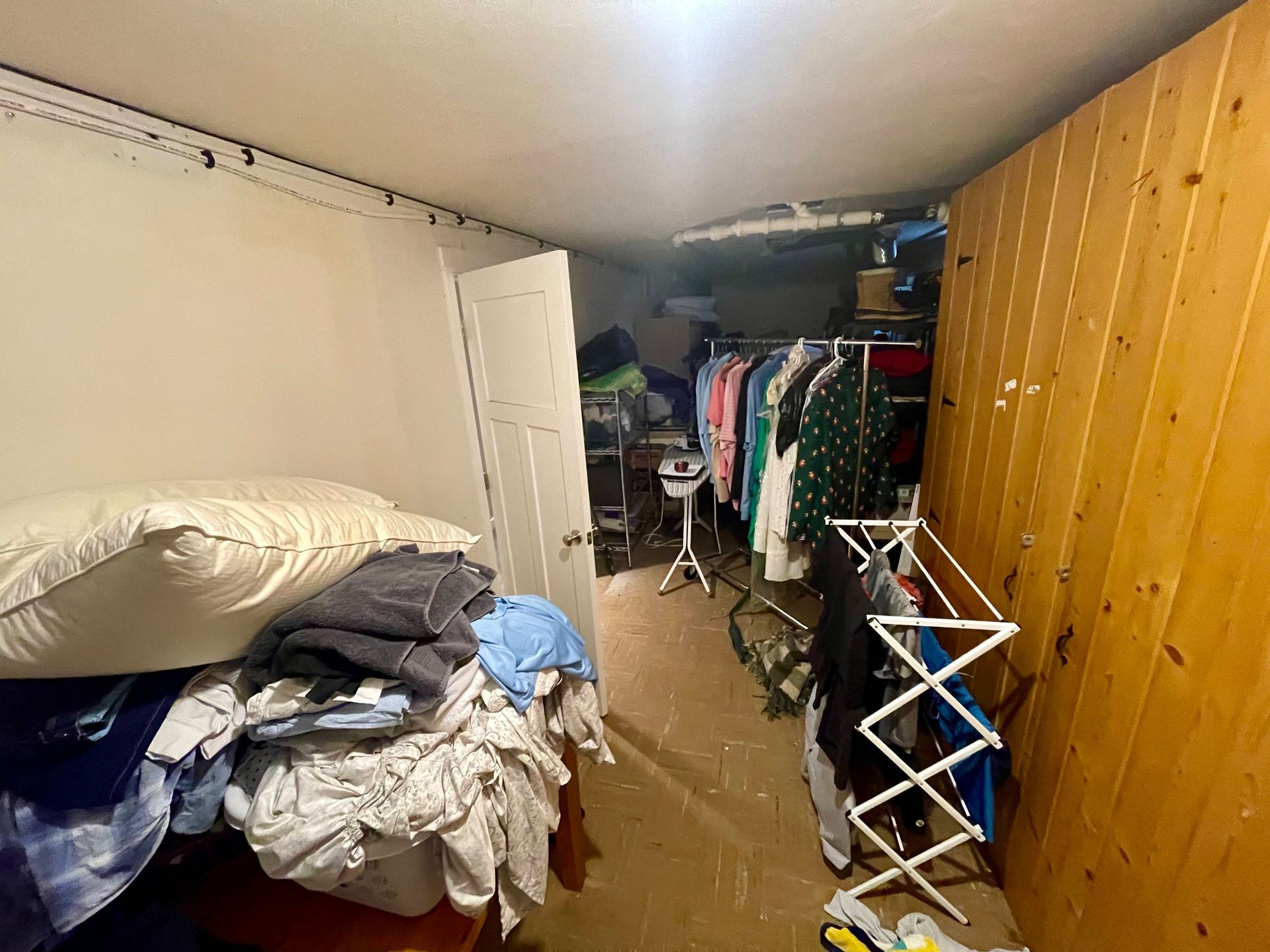 Cluttered, dimly lit storage room. Clothes piled on a bed, hanging rack, and wooden paneling.