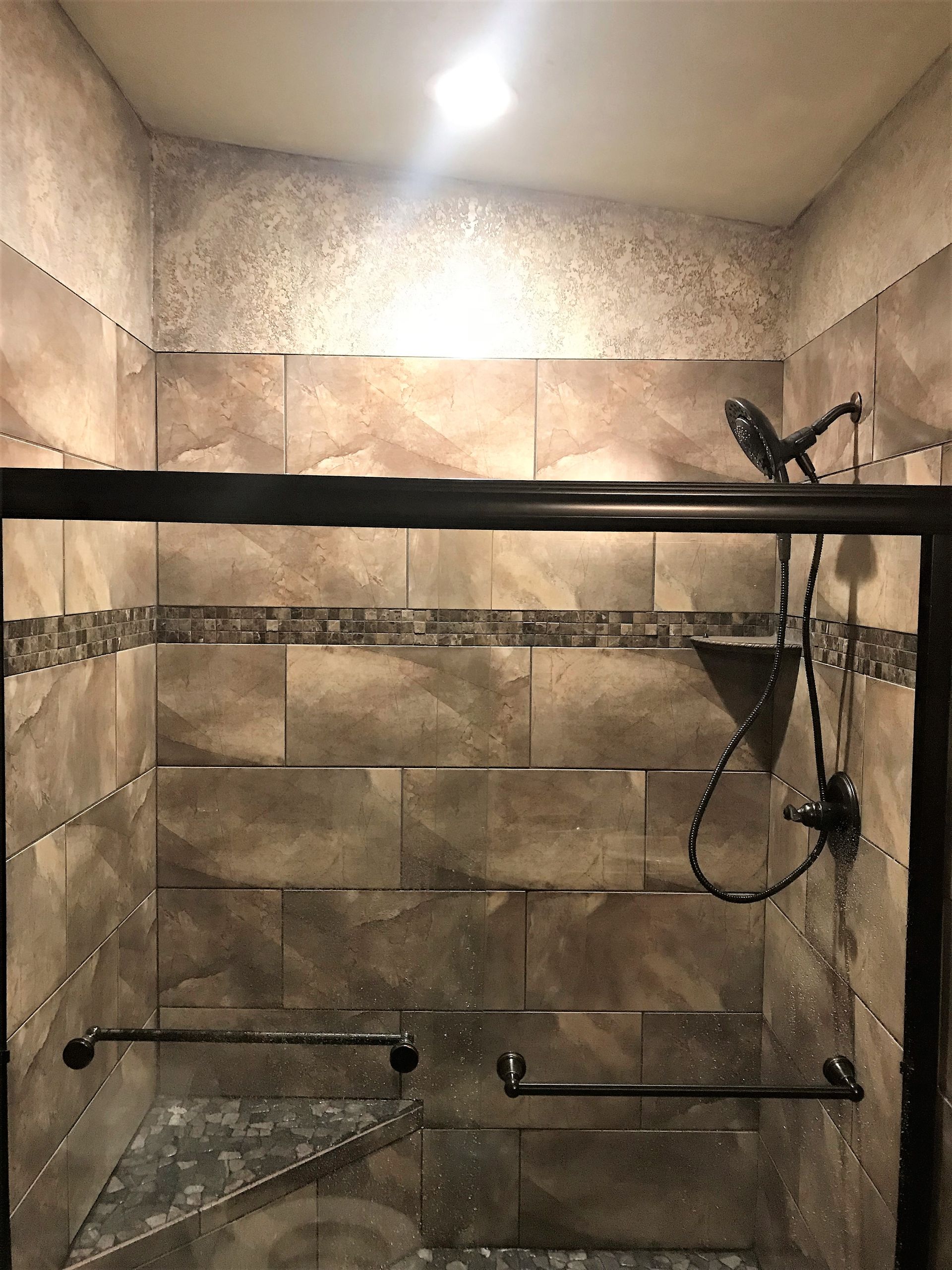 Shower stall with brown tiles, glass door, dark fixtures, and pebble floor.