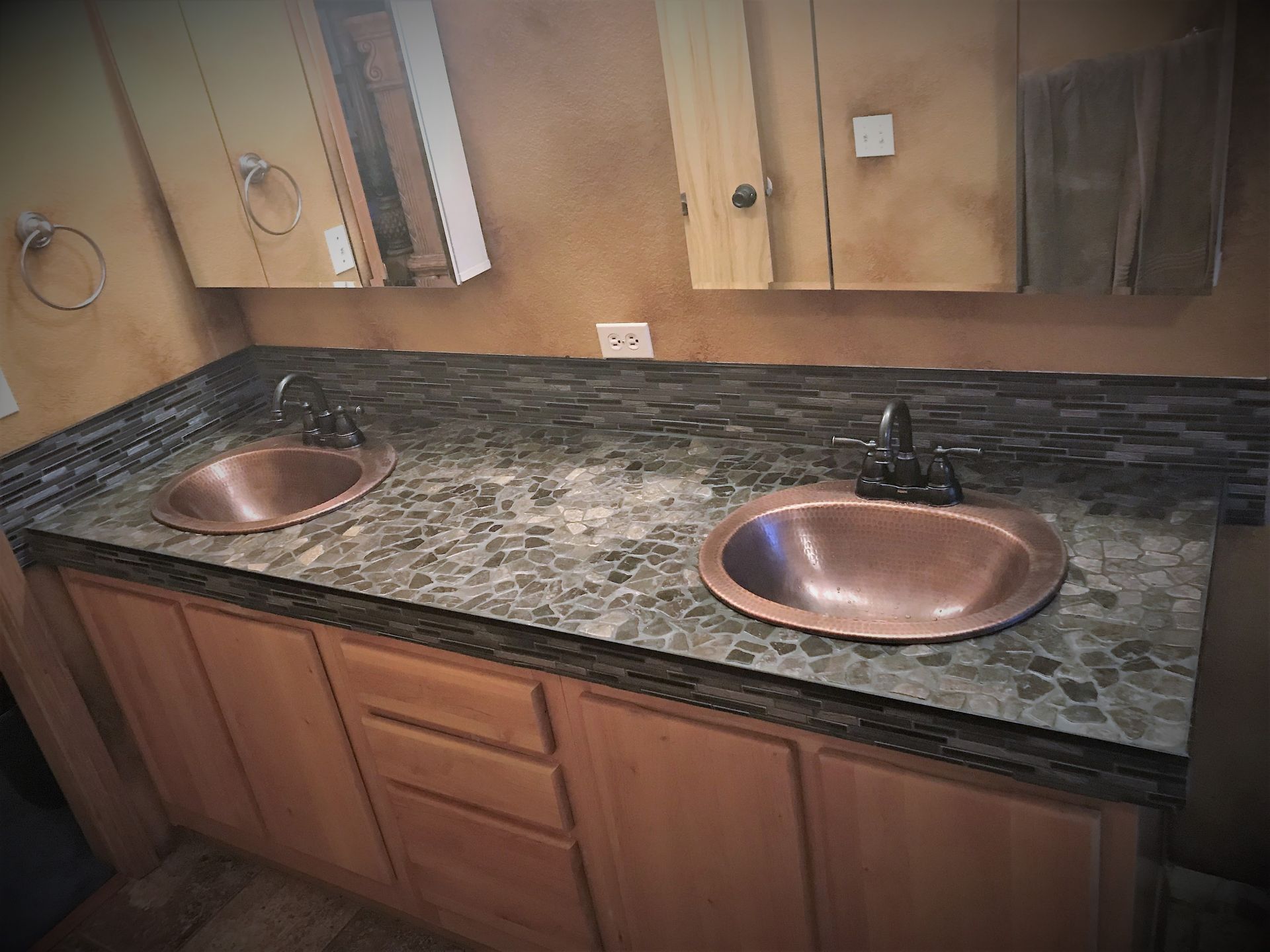 Bathroom vanity with copper sinks, dark countertop, wooden cabinets, and neutral walls.