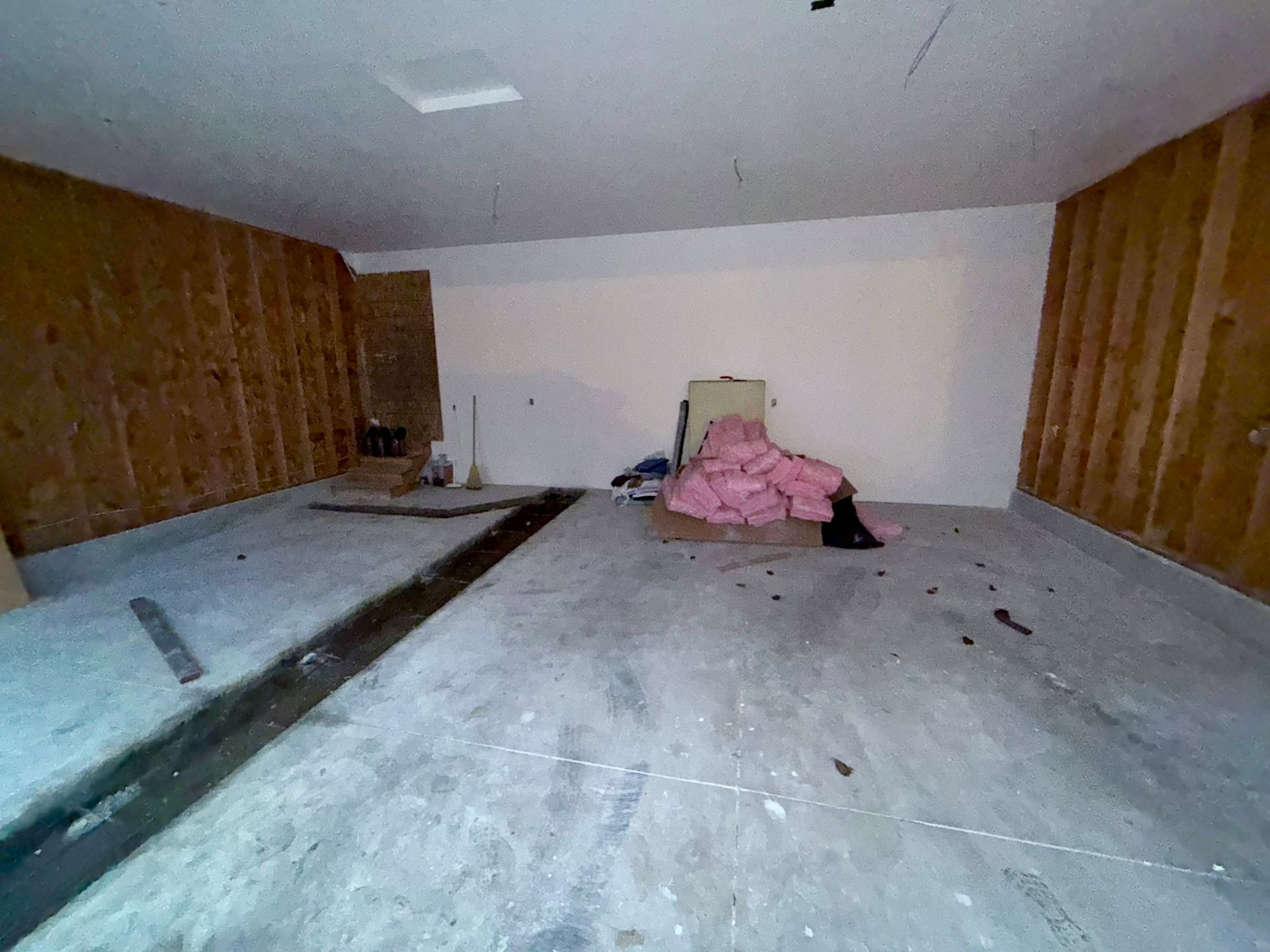 Empty unfinished garage interior with pink insulation, bare plywood walls, and concrete floor.