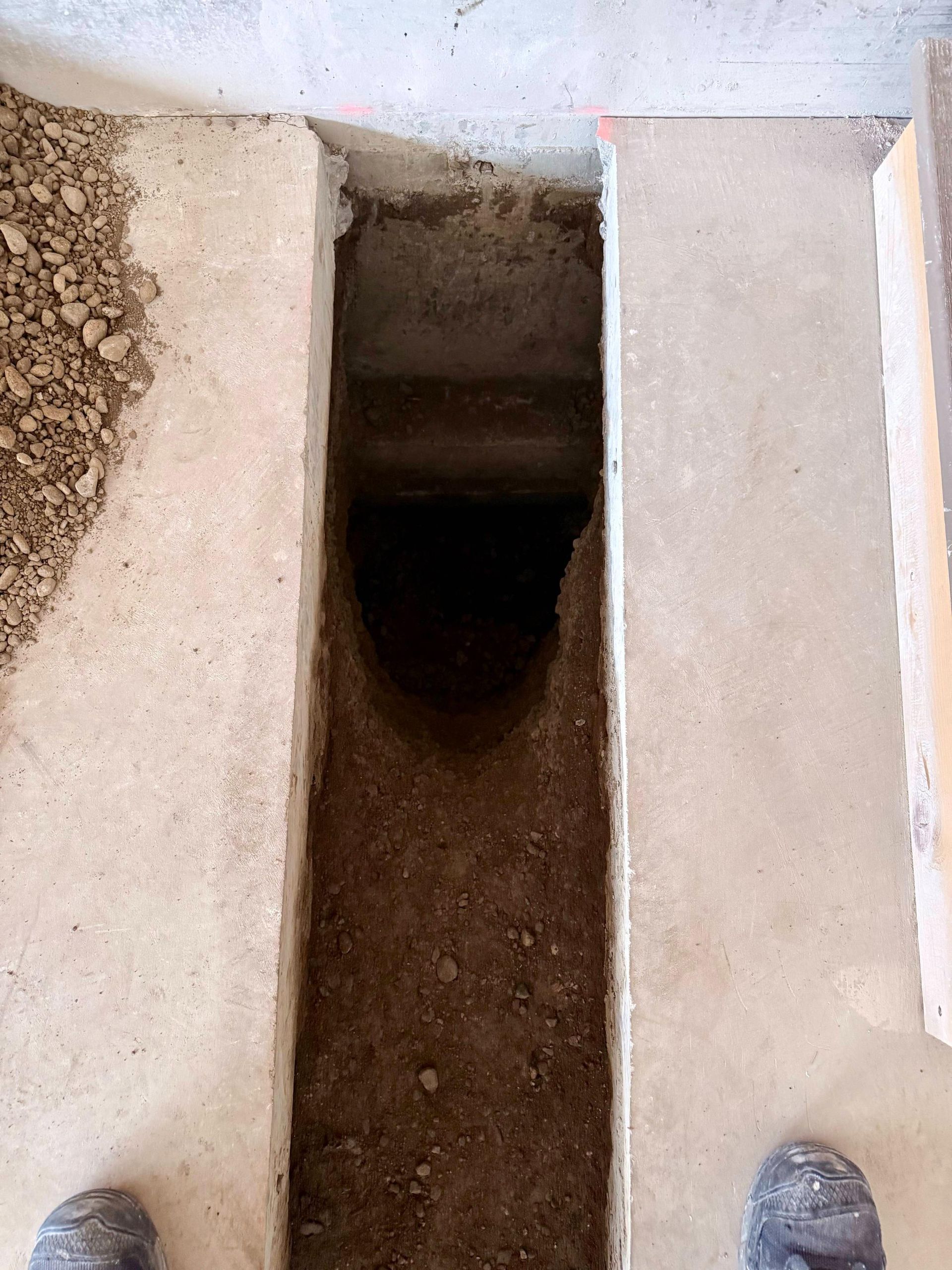 Open trench in concrete floor, filled with dirt; feet visible at bottom.