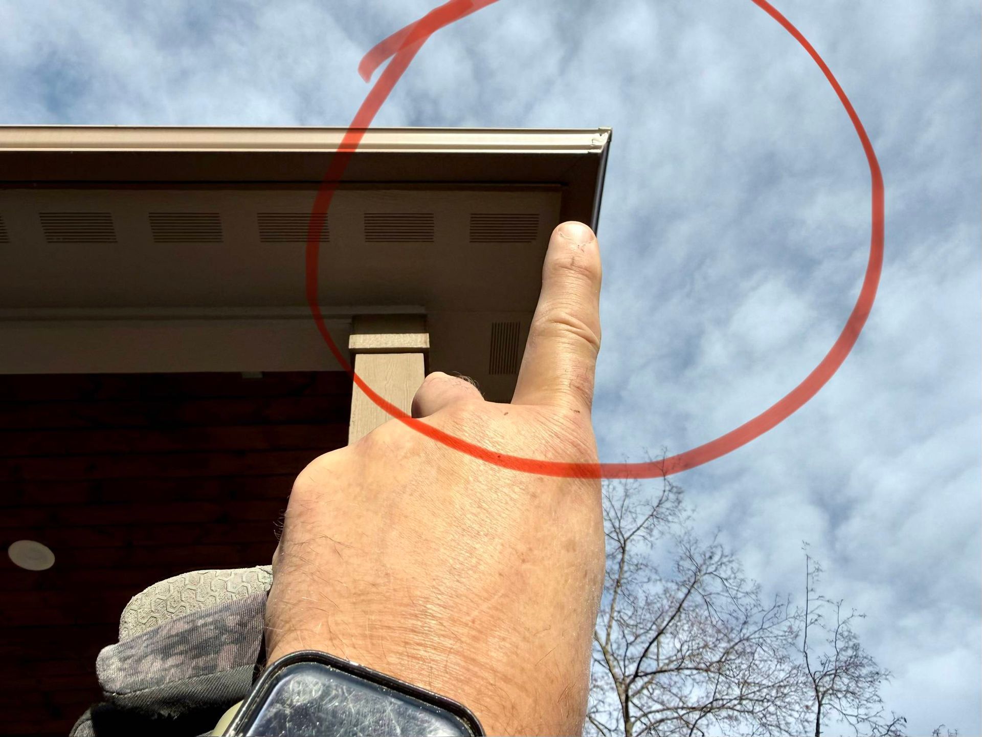 Hand pointing up at a corner of a building with a beige soffit, brown siding, and cloudy sky.