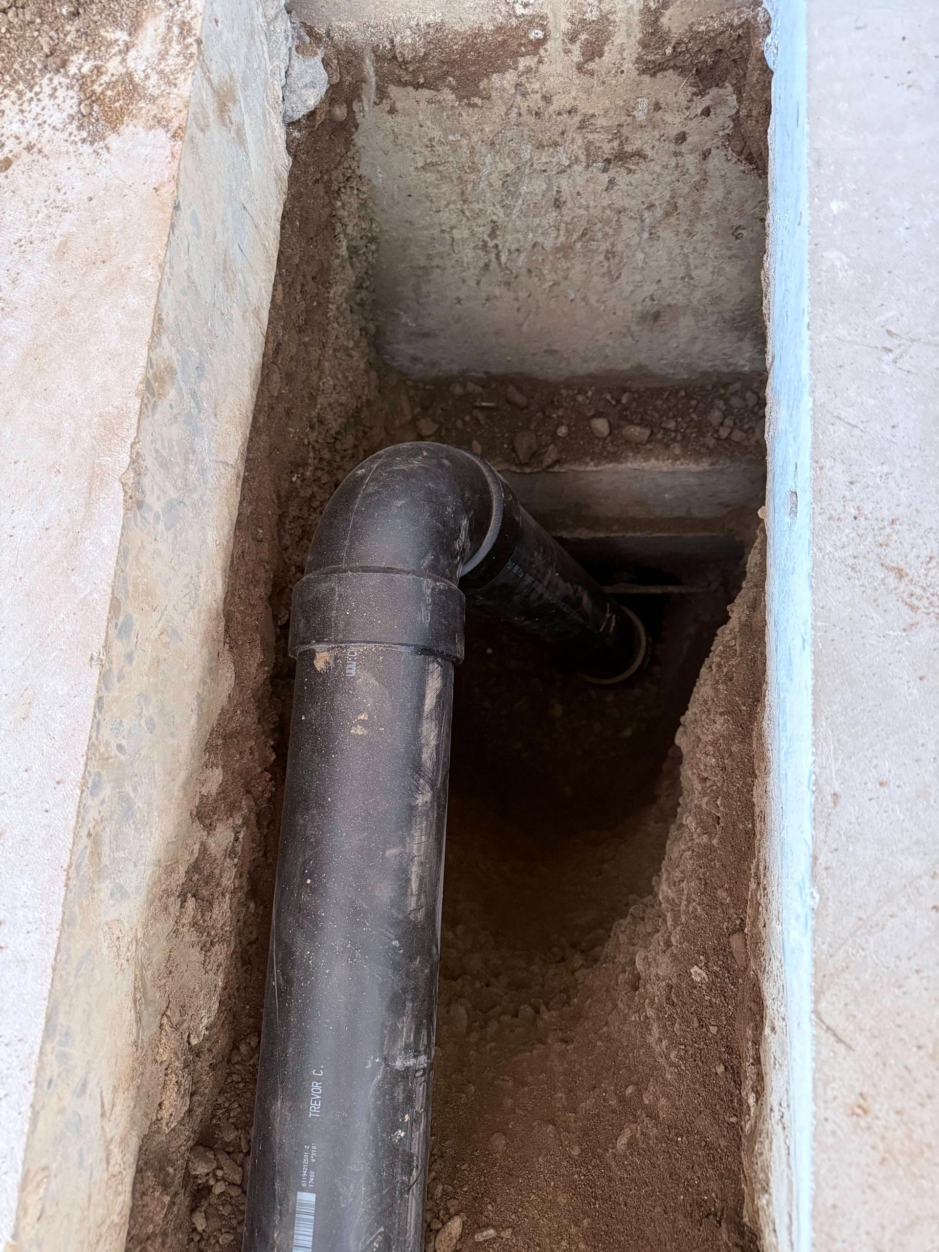 Black pipe with elbow fitting in a concrete trench filled with dirt.