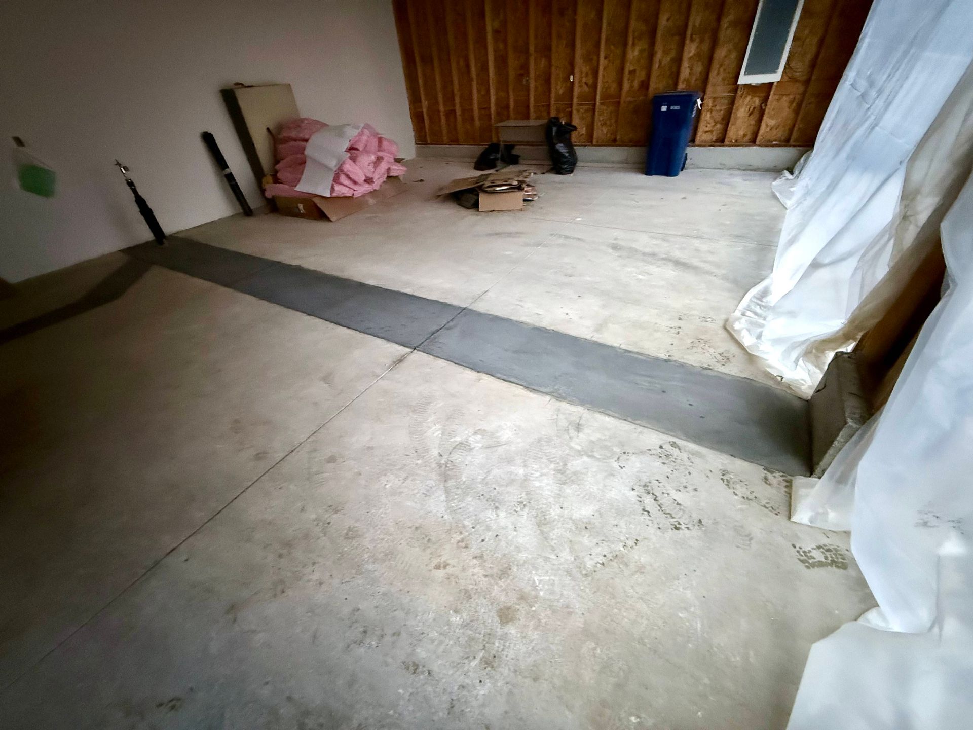 Garage interior with concrete floor, insulation, trash can, and lumber framing the wall.