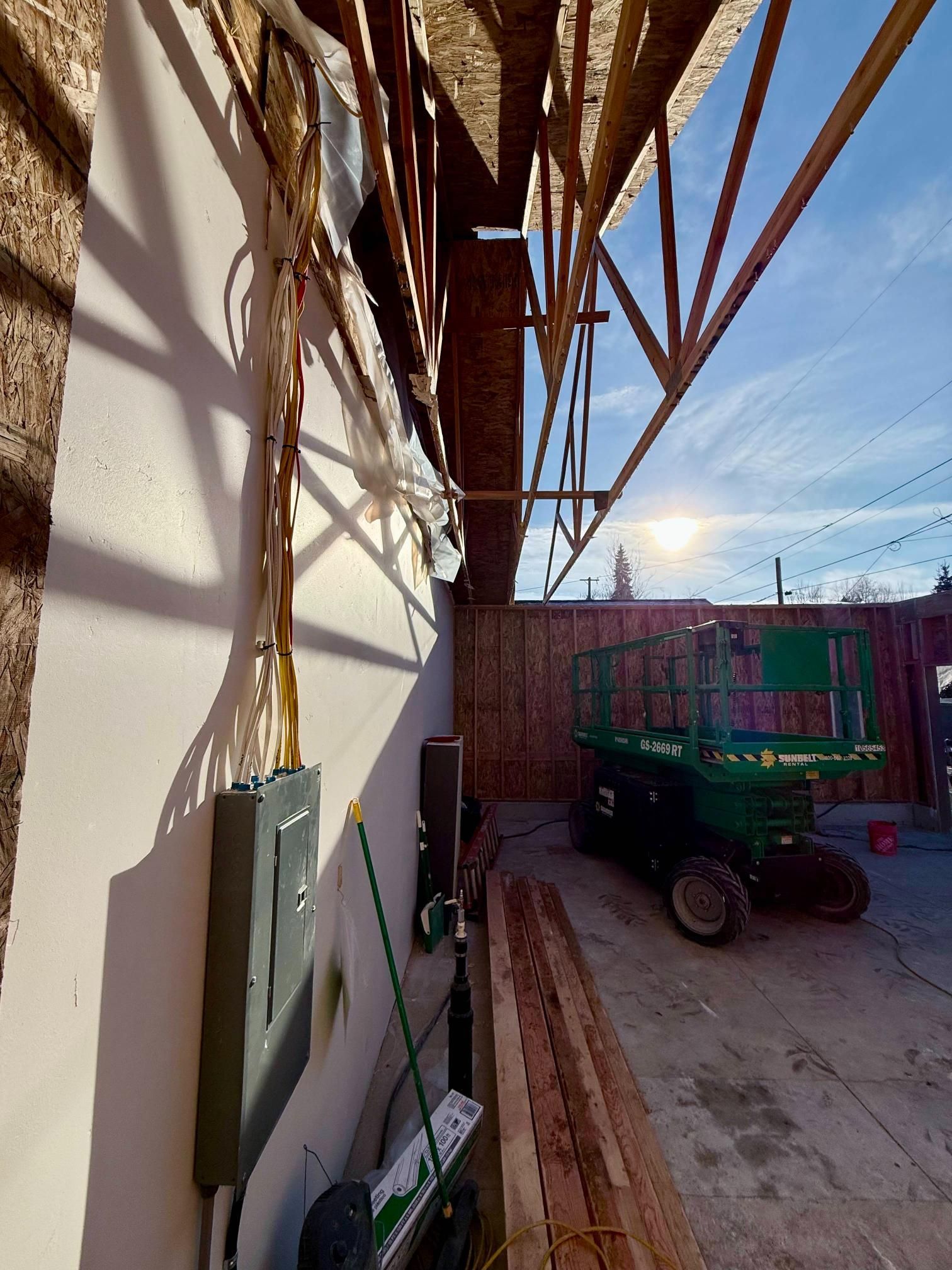 Construction site: electrical panel on a white wall, wooden beams overhead, lift, lumber, and a sunny sky.