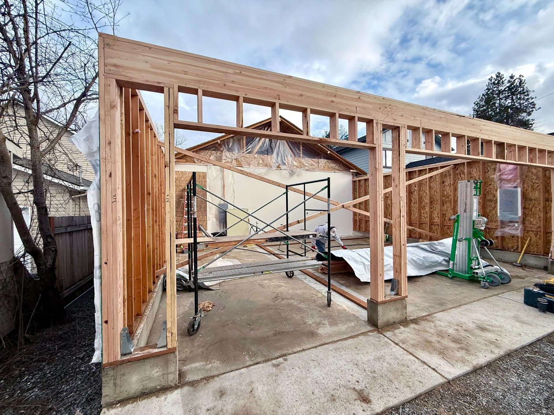 Framing of a garage extension in progress; wood beams, scaffolding, overcast sky.