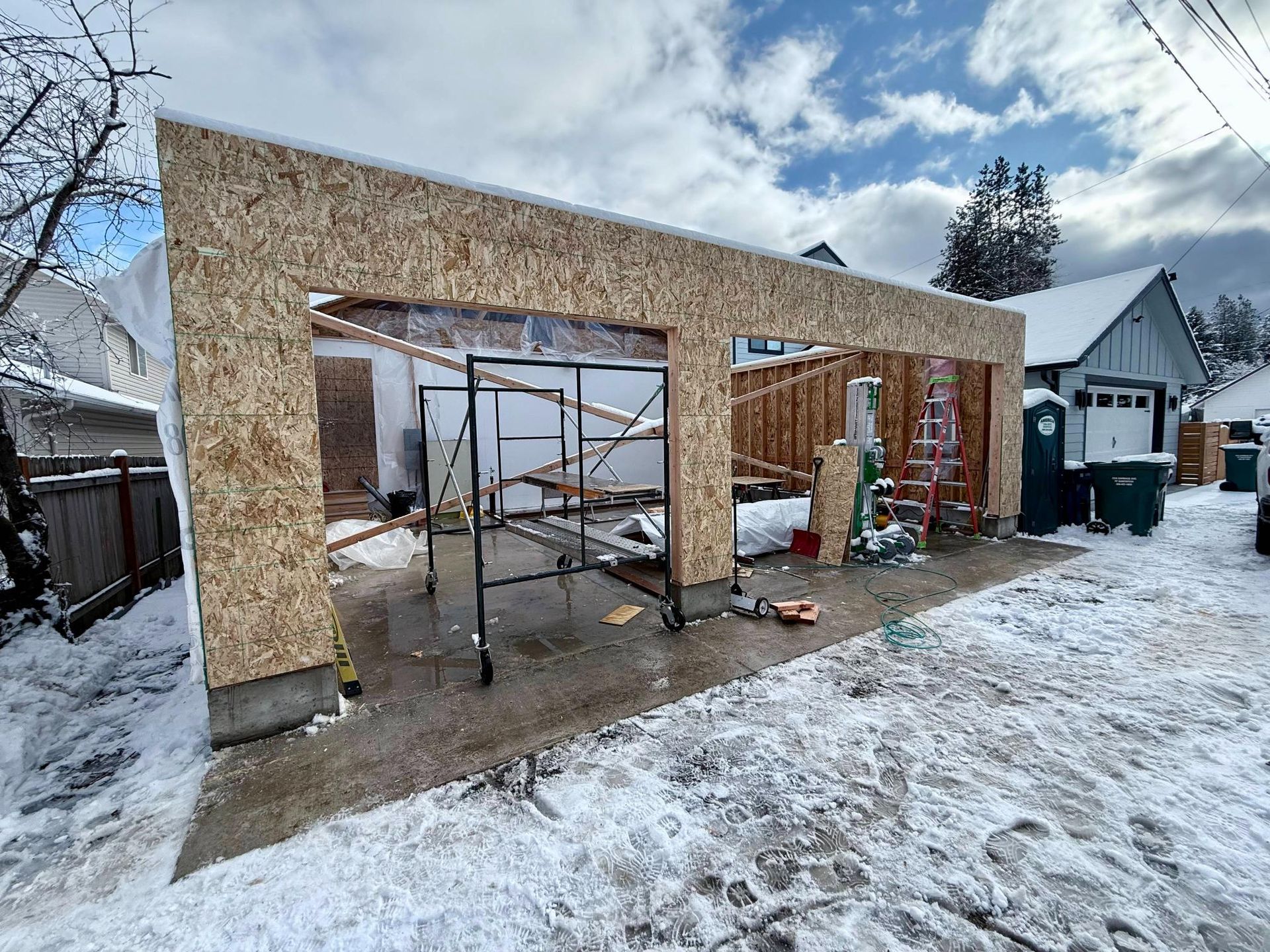 Garage construction in snowy conditions: OSB walls framing a concrete slab, scaffolding, cloudy sky.