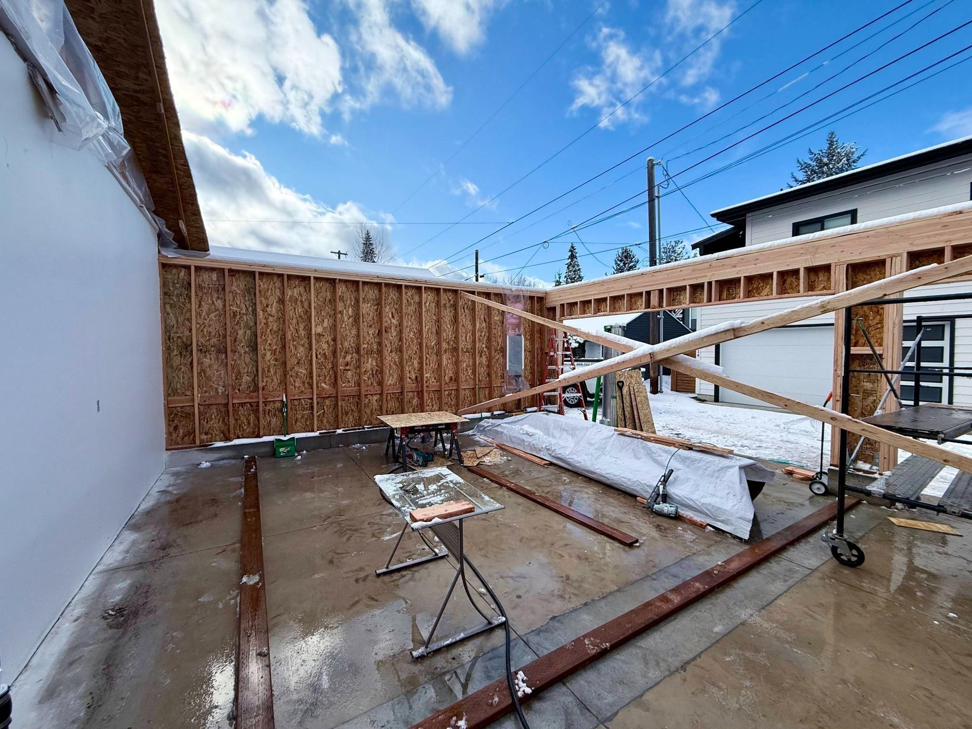 Construction site: wood frame structure, OSB walls, snow-covered ground, sunny sky.