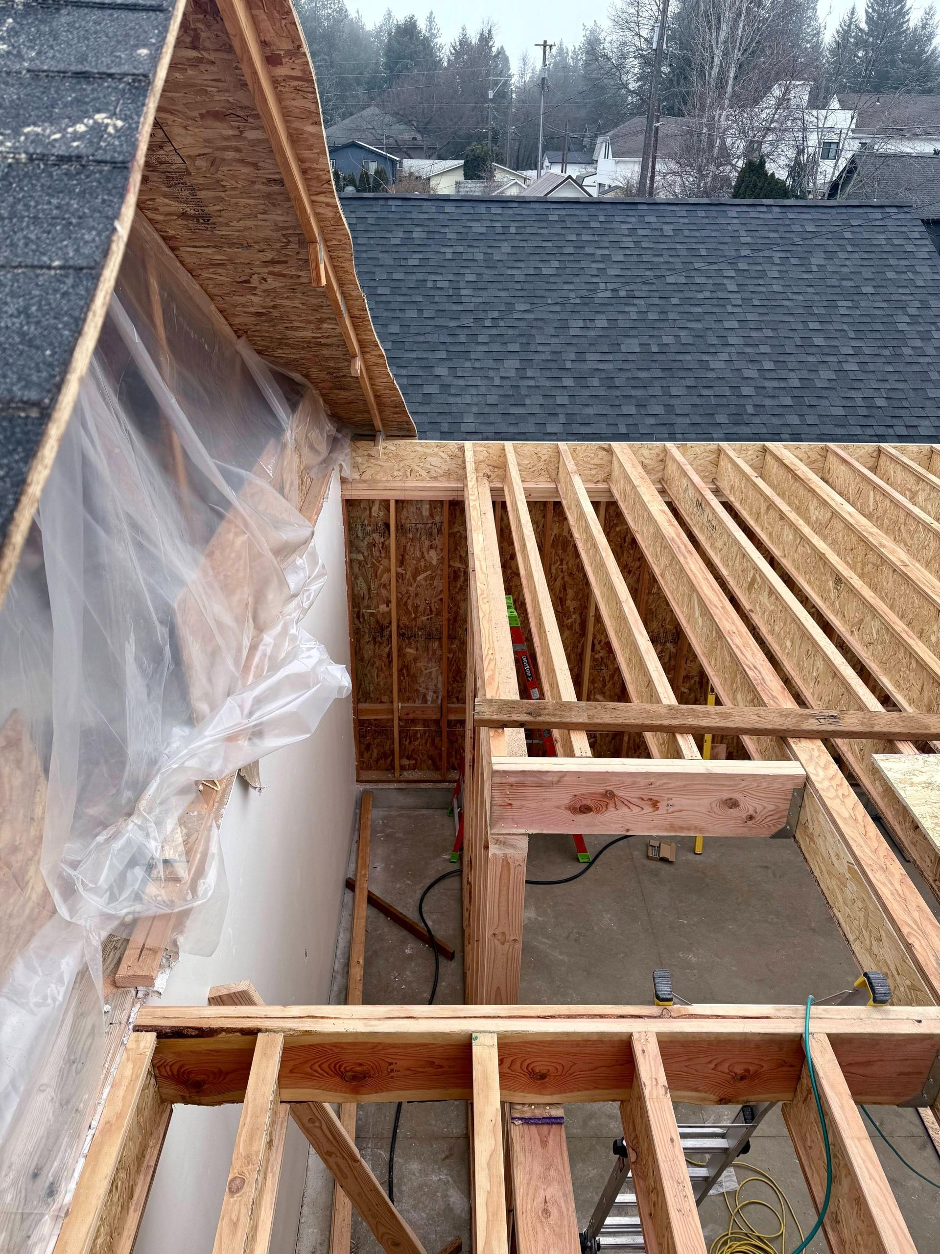 Wood framing and roofing in progress; construction site with tarp-covered wall, visible rafters, and dark shingles.