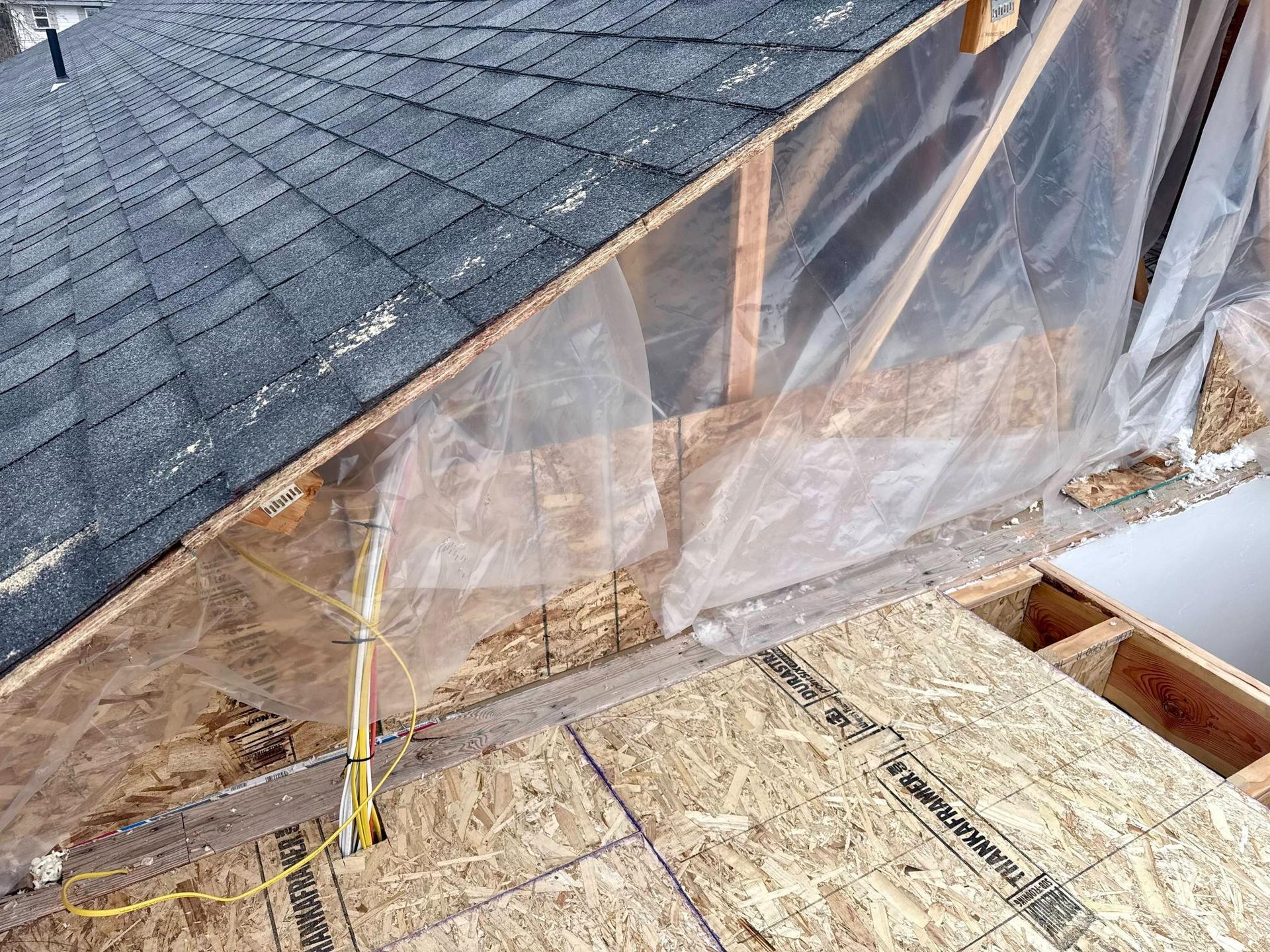 Interior view of an unfinished attic space with roofing shingles, exposed wooden beams, and a plastic sheet.