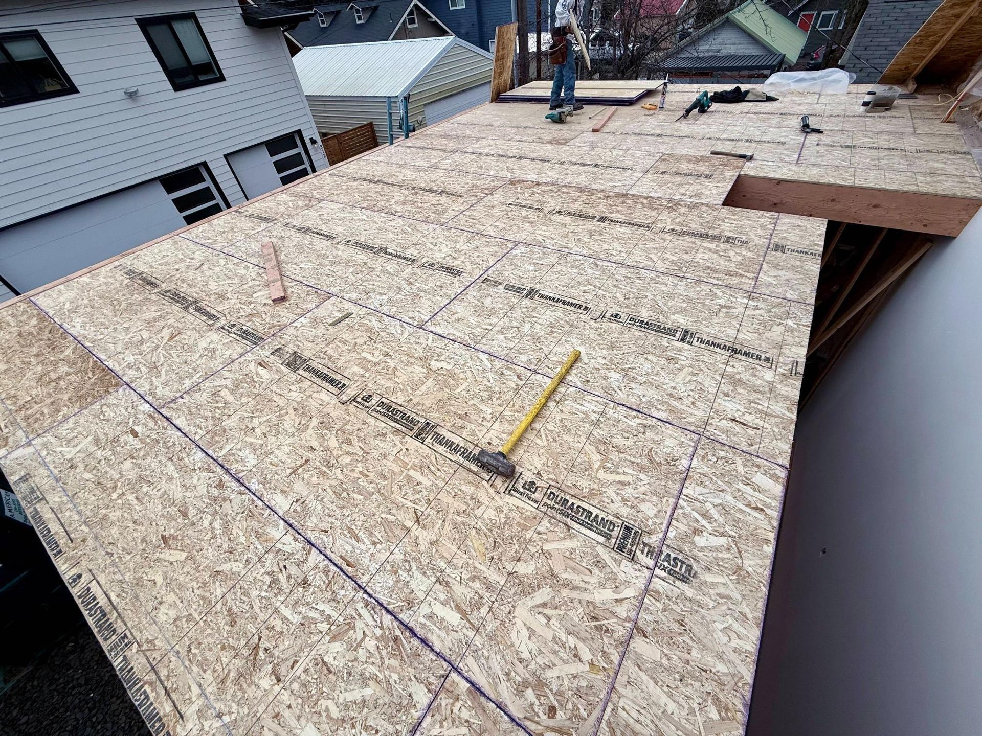 Rooftop construction: OSB sheathing on roof frame, tools scattered, person working in background.