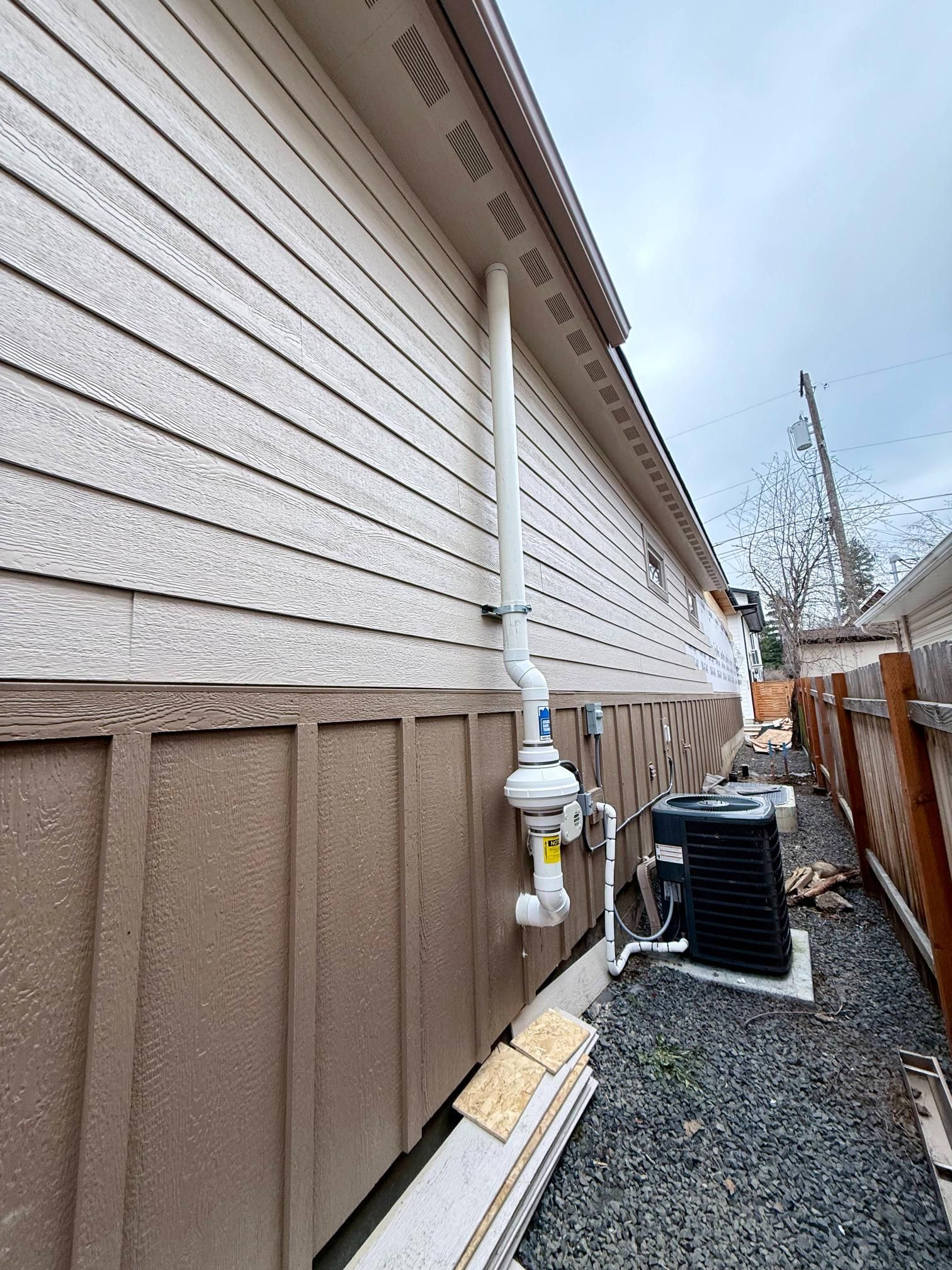 Exterior of a building with a white gas pipe and air conditioning unit along the side. Brown siding and a gravel pathway.