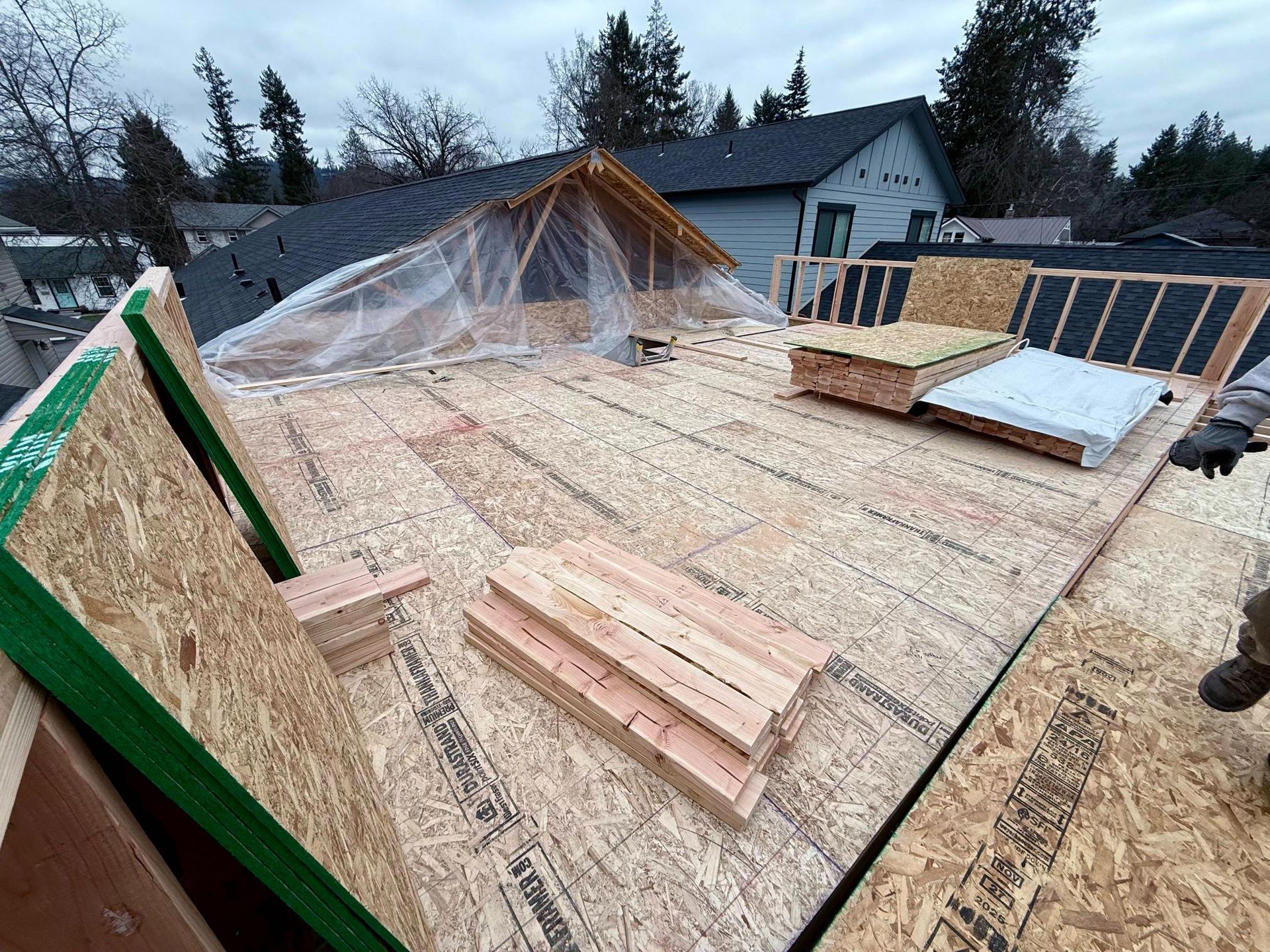 Rooftop construction: plywood and lumber laid out on a roof, tarp over frame, cloudy sky.