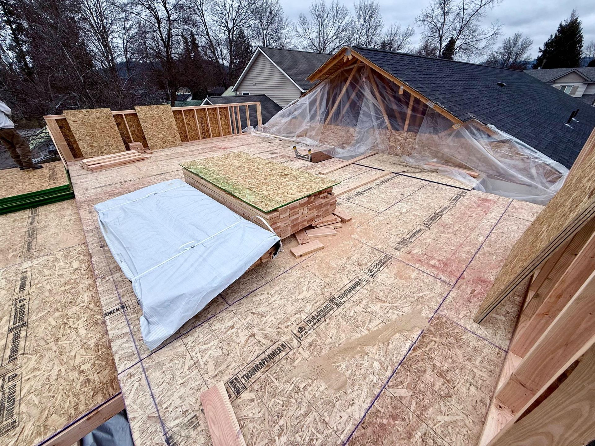 Rooftop construction: plywood deck with exposed framing, bundles of plywood, plastic tarp covering part of the roof.