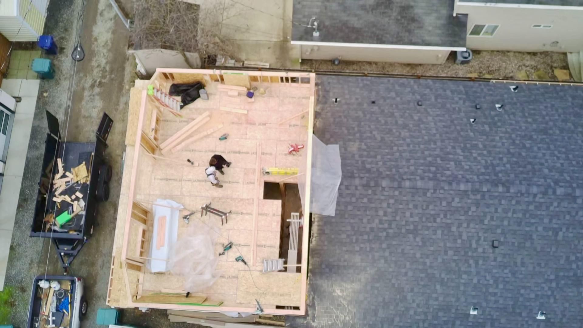 Construction site from above: A partially built wooden structure with a worker. Next to a roof with dark shingles.