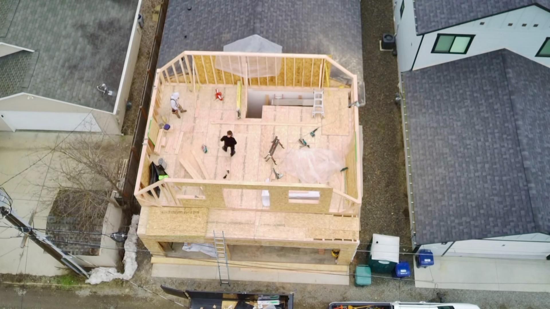 Aerial view of a two-story house under construction, workers on the wooden structure, surrounded by other buildings.