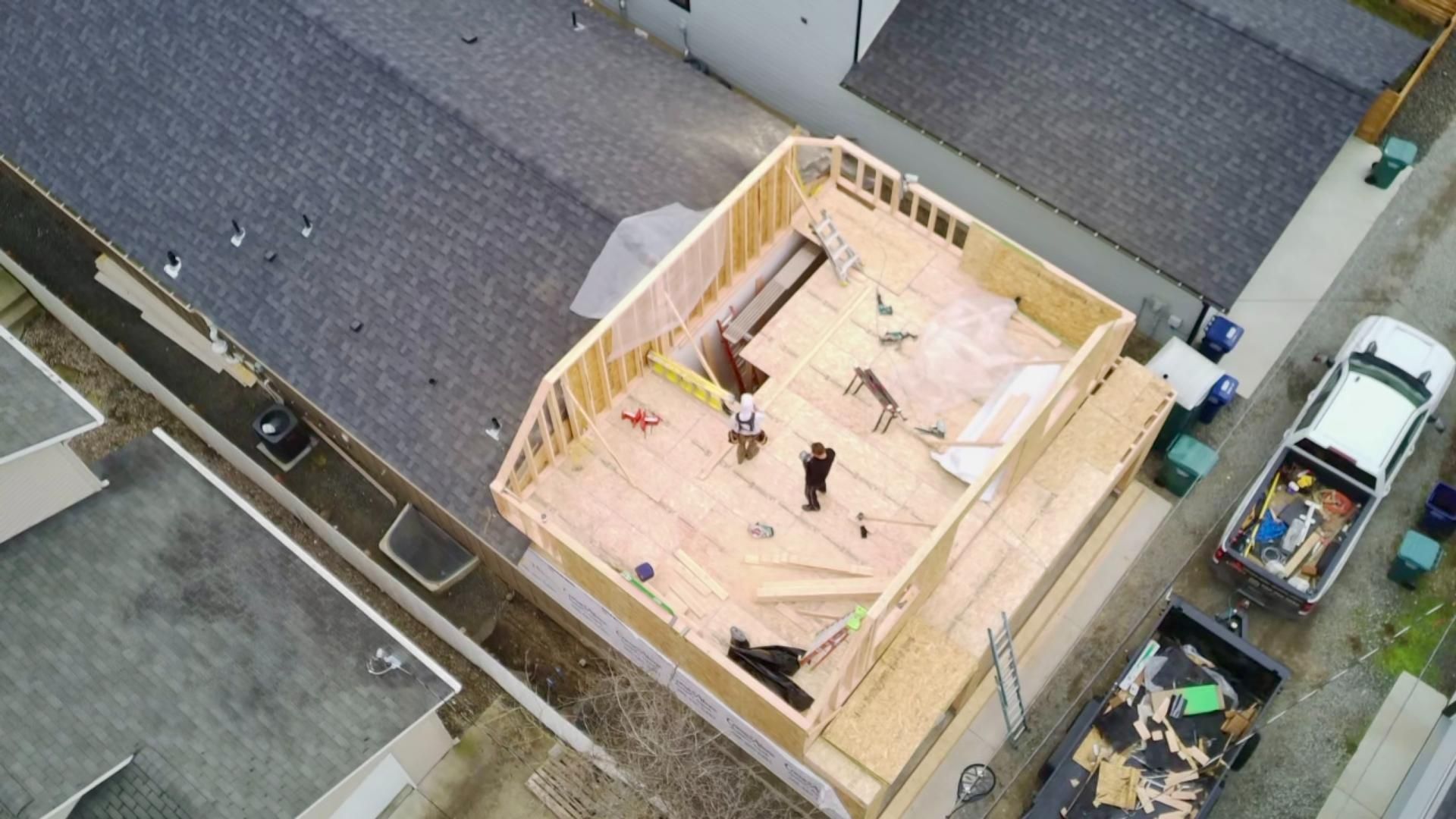 Overhead view of a building under construction, wooden frame with workers, next to a street with cars and neighboring houses.