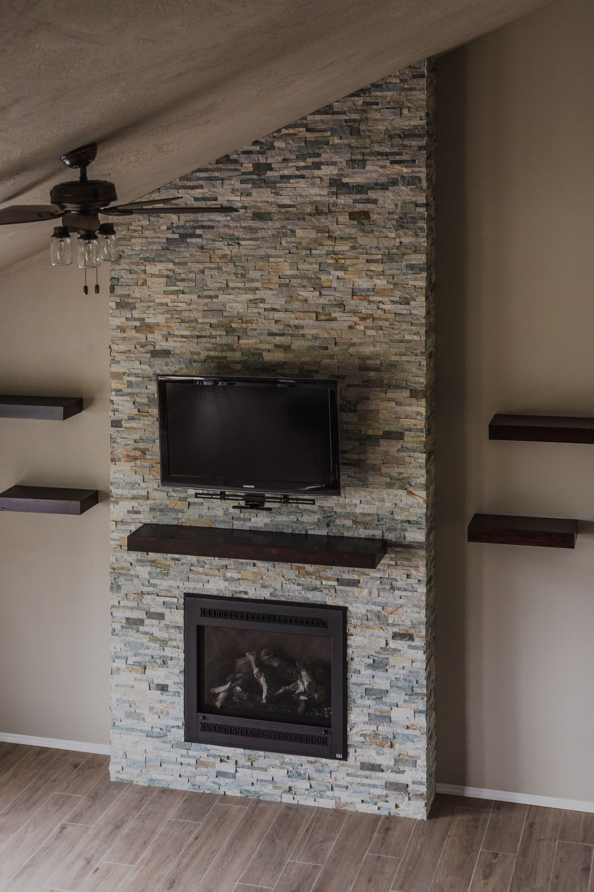 Stone fireplace wall with mounted TV, fireplace, and floating shelves in a room with an angled ceiling.