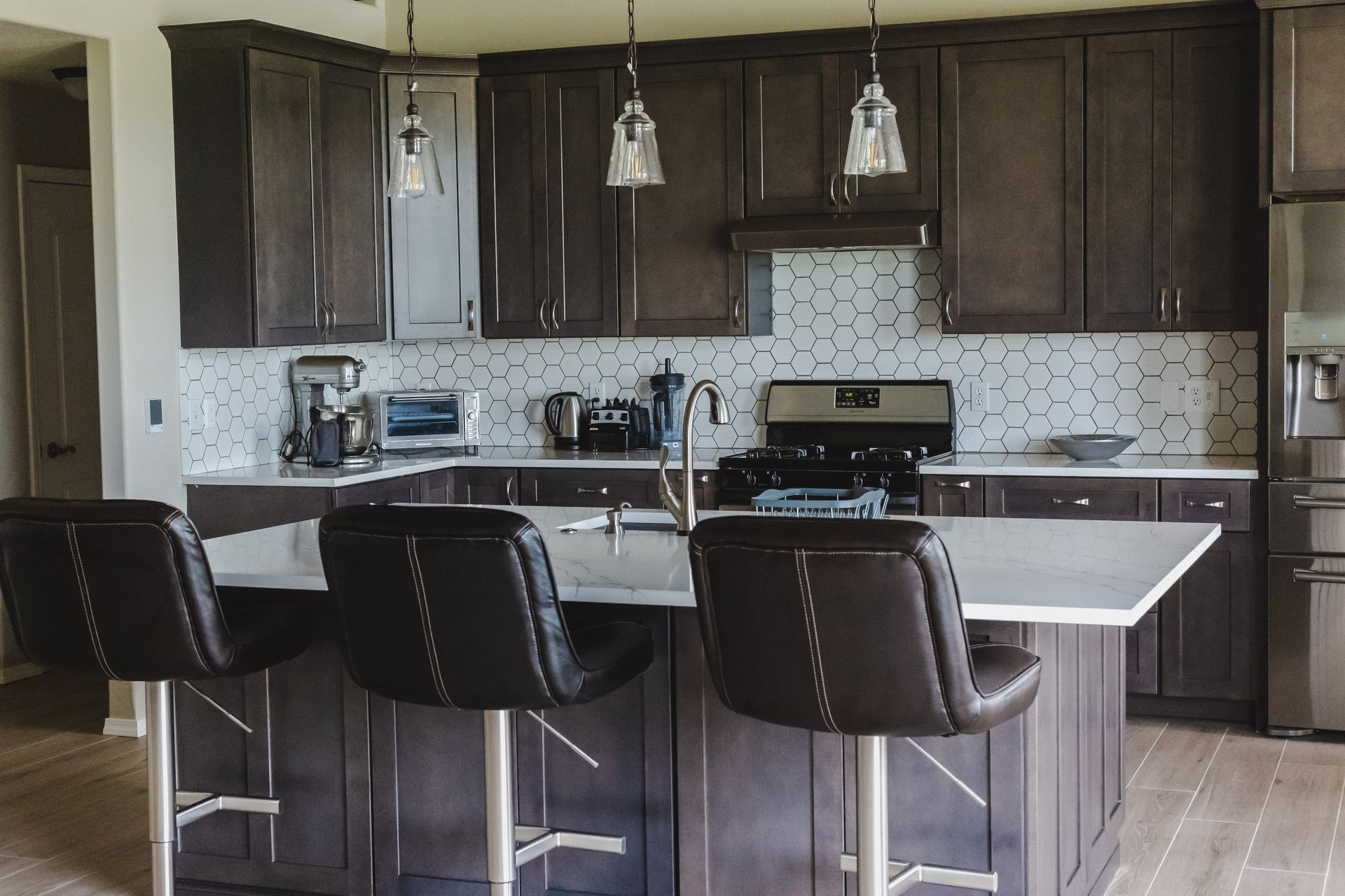 Kitchen with dark wood cabinets, island with bar stools, stainless steel appliances, and white backsplash.