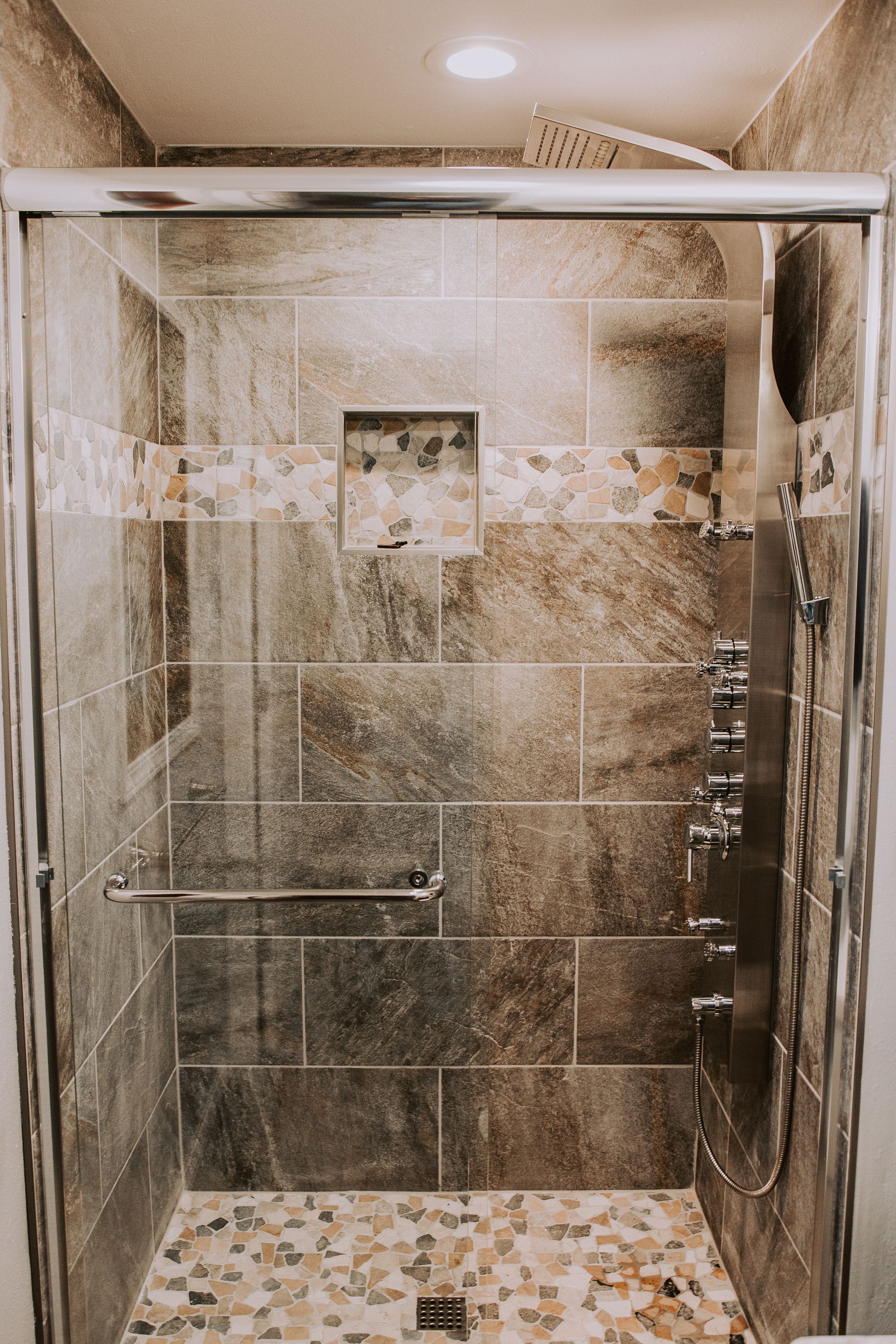 Shower with brown tiled walls, stone floor, glass door, and multiple showerheads and controls.