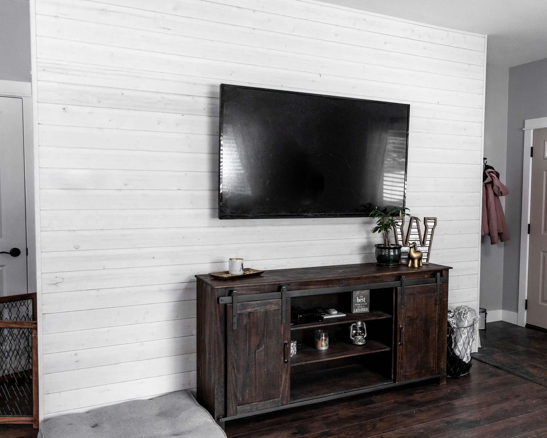 White shiplap wall with large TV above a dark wood cabinet. A door, bench, and entry are visible.