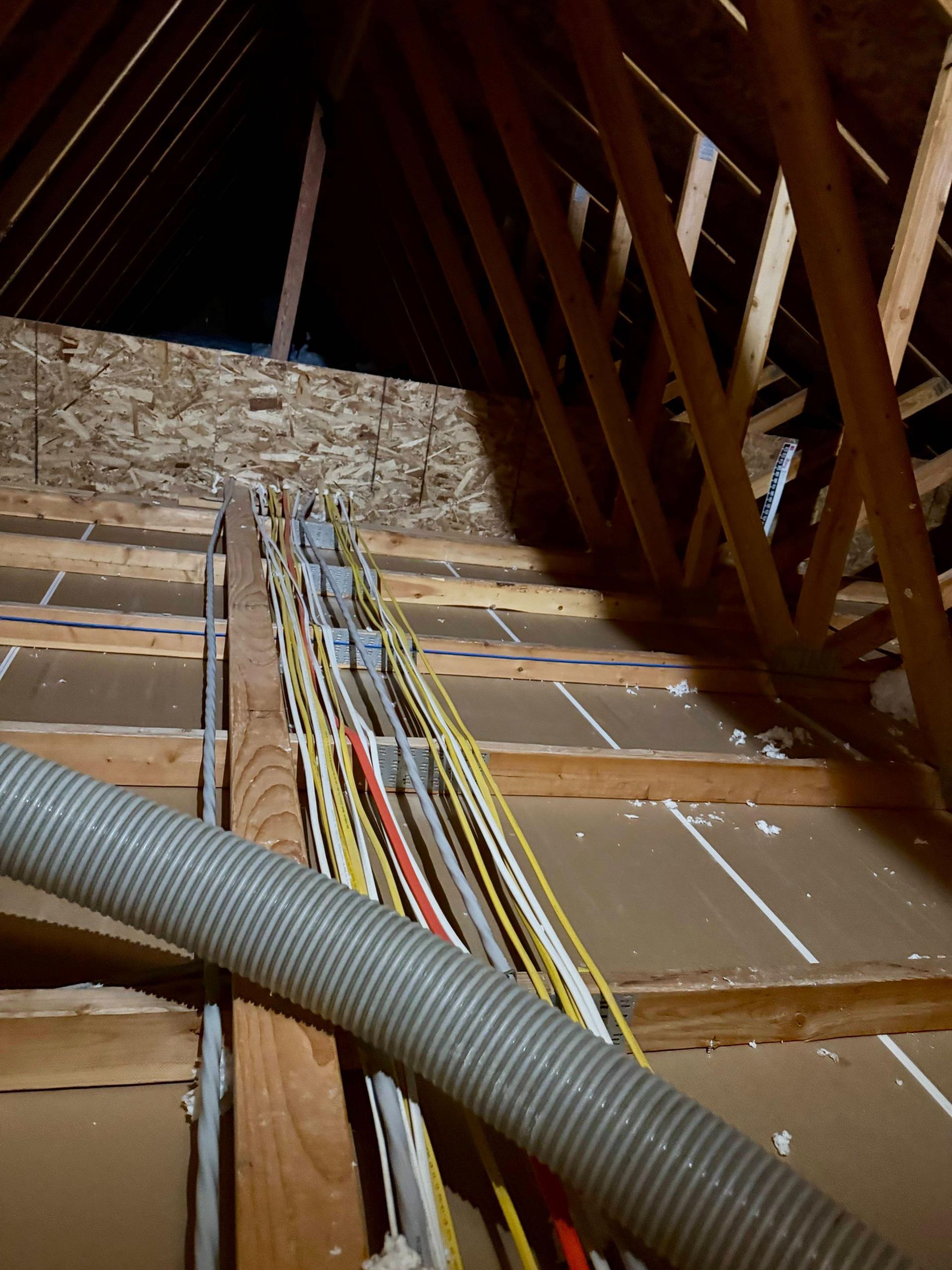Electrical wiring and a flexible gray tube run through a dark attic space, supported by wooden beams.