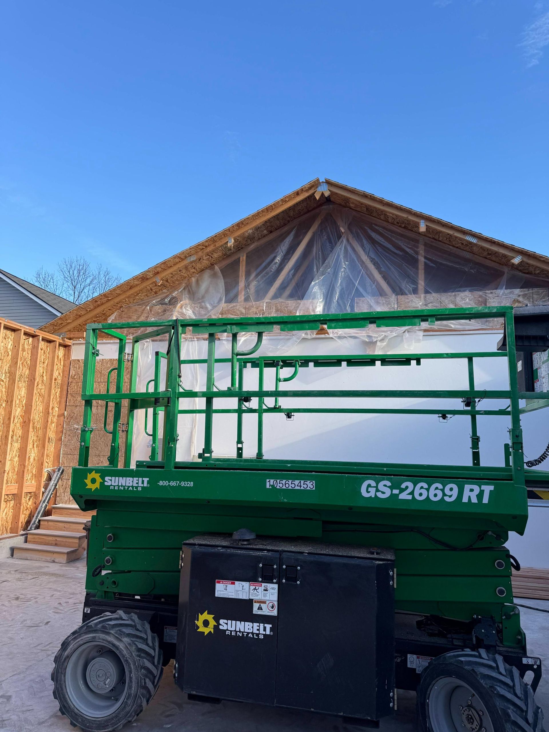 Green scissor lift in front of a building under construction, blue sky.