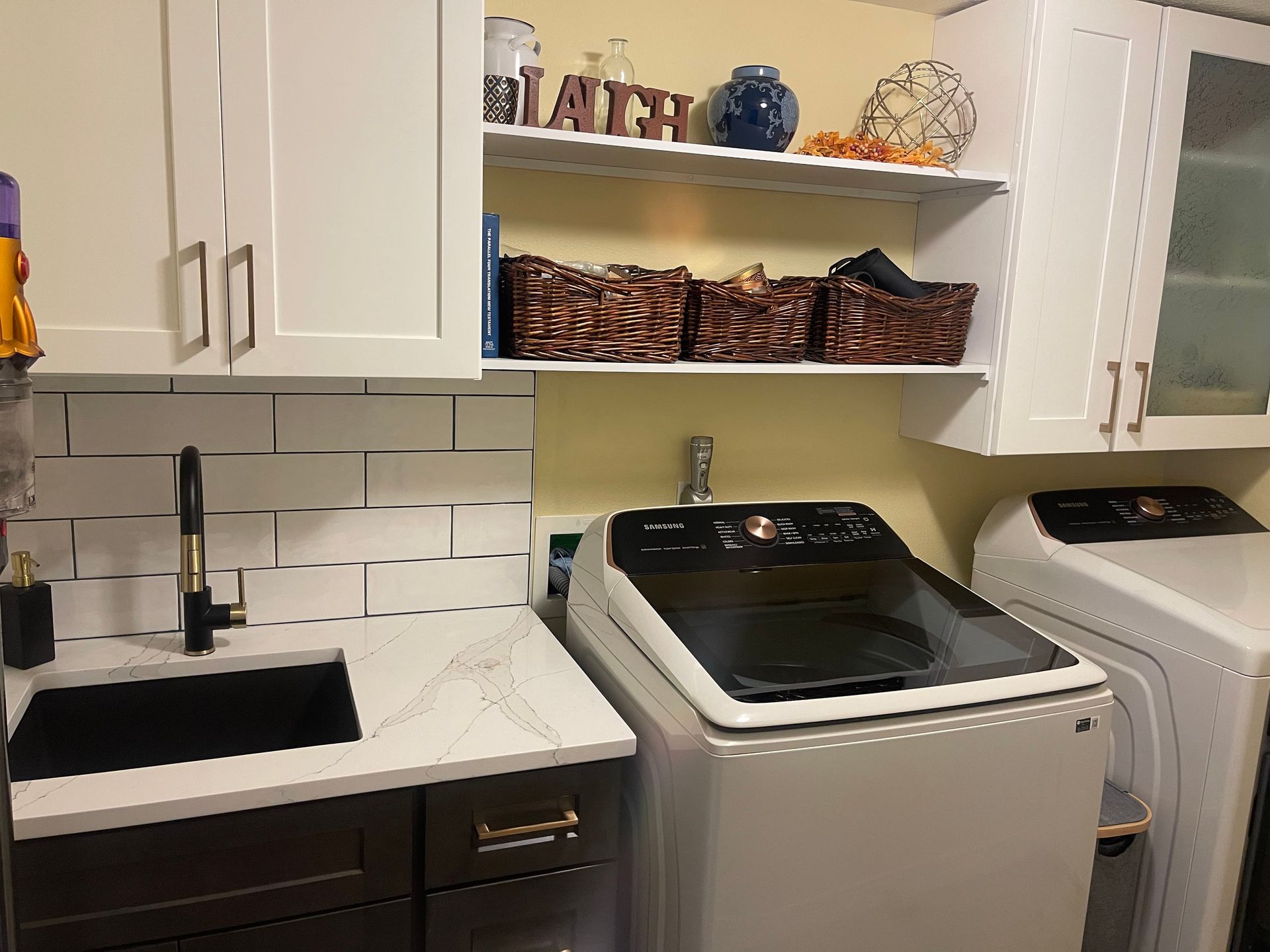 Laundry room with white cabinets, black sink and faucet, and washer/dryer. Shelves with baskets and decorations.
