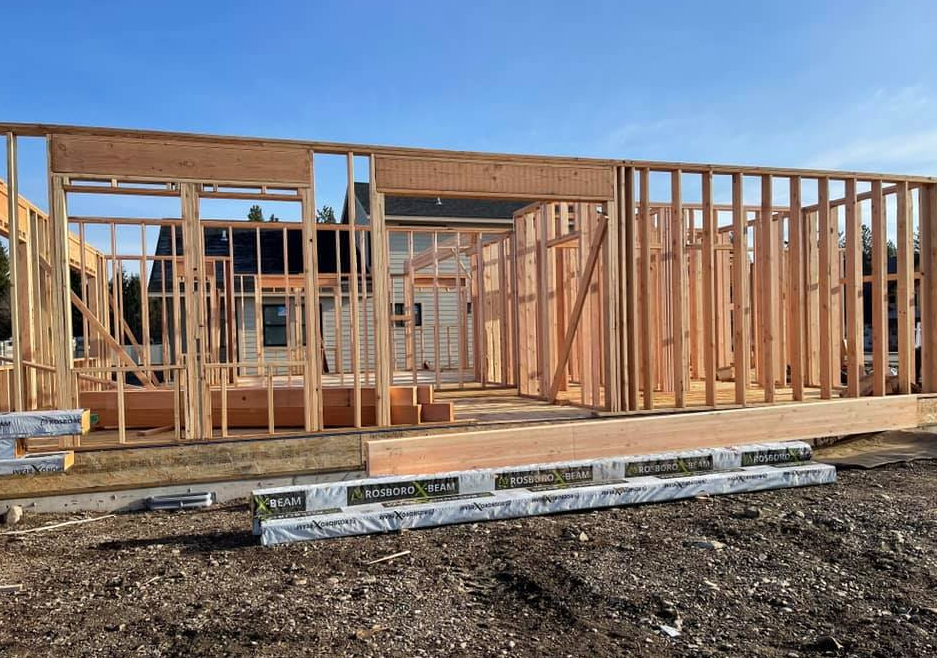 Framing of a wood-framed house under construction on a dirt lot, with the existing home in the background.