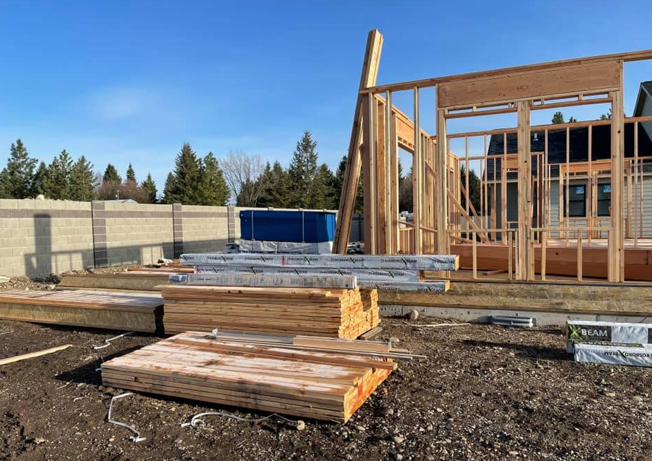 Construction site: wooden framework of a house being built, with stacks of lumber in front.