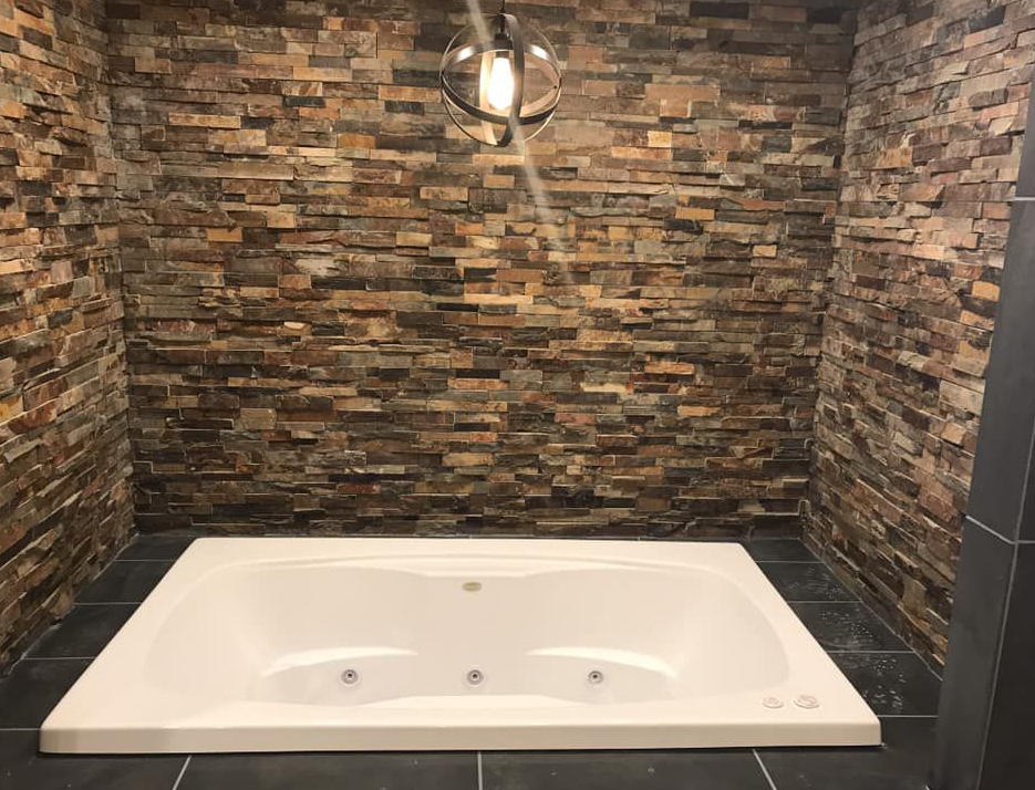 White jacuzzi tub surrounded by stone walls, black tiled floor, and a pendant light.