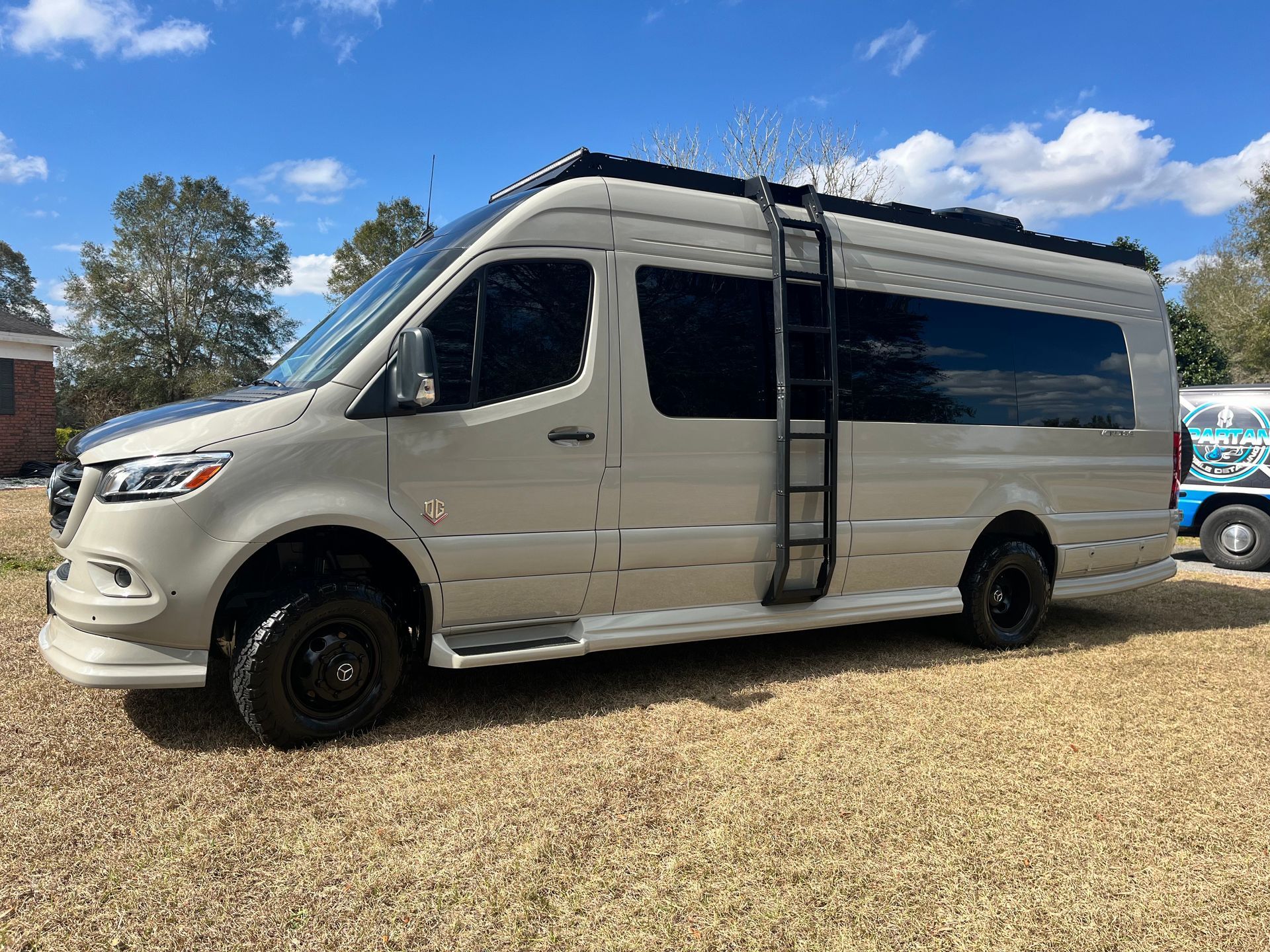 A white van with a ladder on the side is parked in a gravel lot.