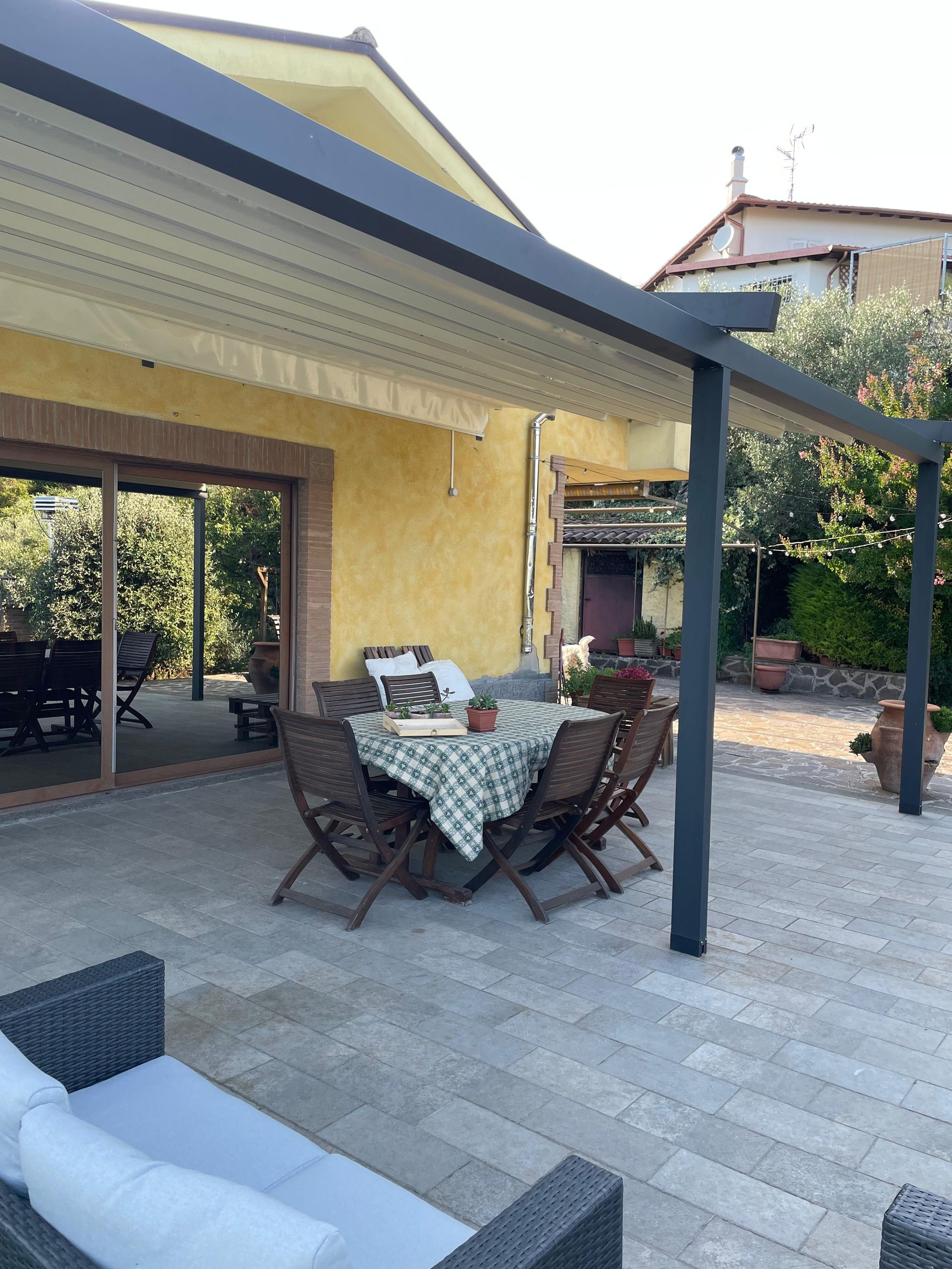 Patio con tavolo, sedie e pergolato di fronte a un edificio giallo con zona pranzo all'aperto visibile.