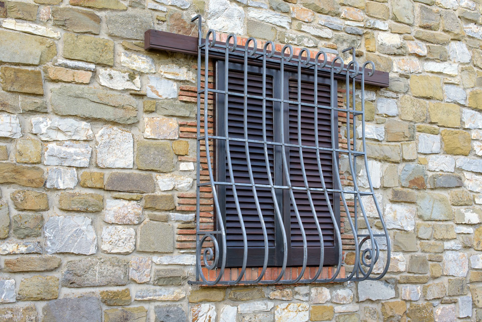Finestra con persiane in legno scuro dietro grate in ferro battuto decorate inserite in un muro di pietra.