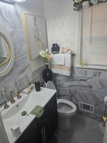 Bathroom with gray marble tile, gold accents, artwork, flowers, and a white vanity.