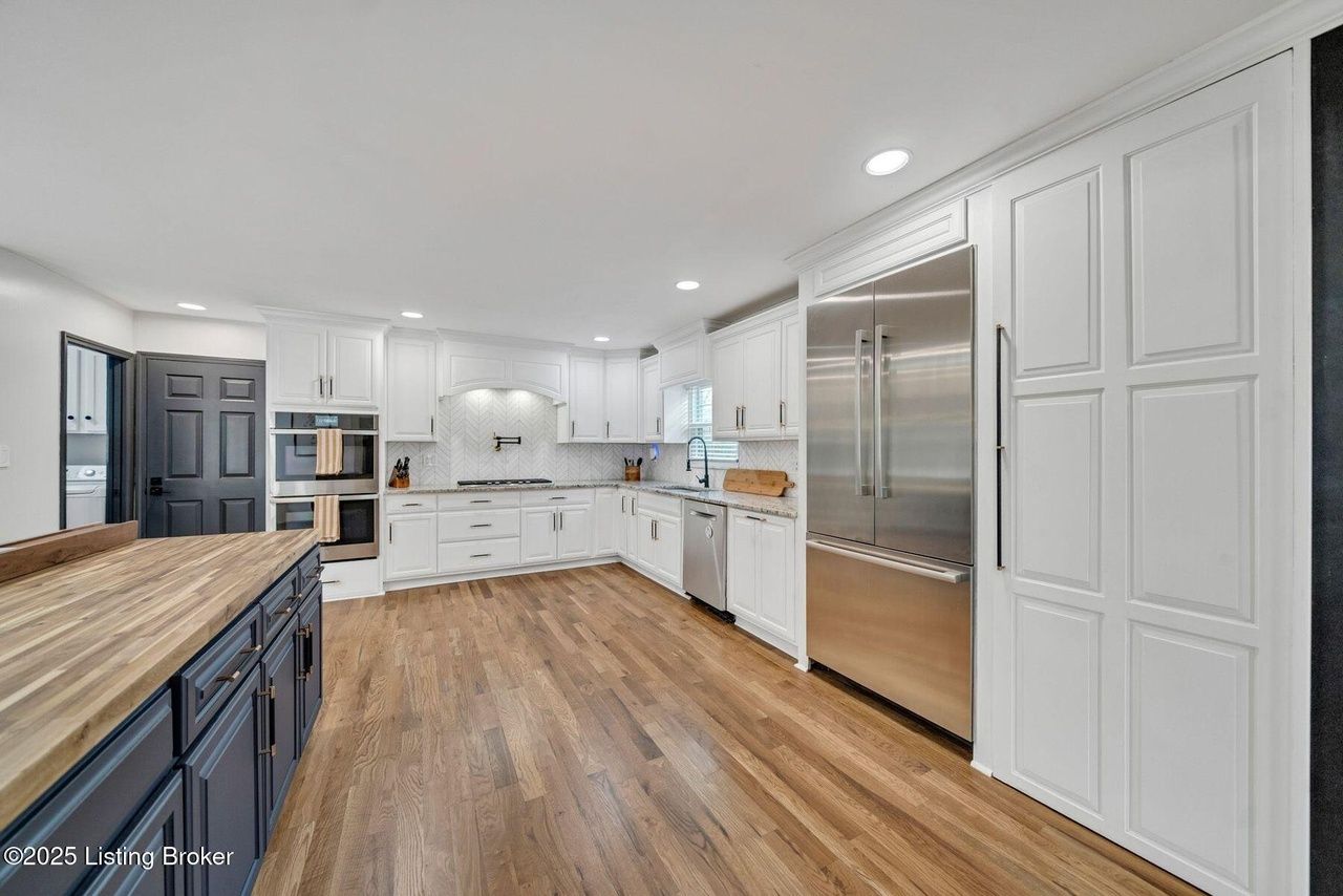 Spacious white kitchen with stainless steel fridge and wooden floors. Island with a blue base and wooden countertop.
