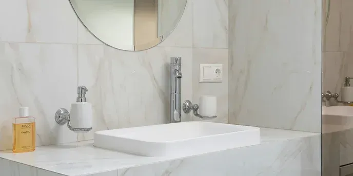 White bathroom sink with chrome faucet, soap dispenser, and matching accessories, set against marble walls.