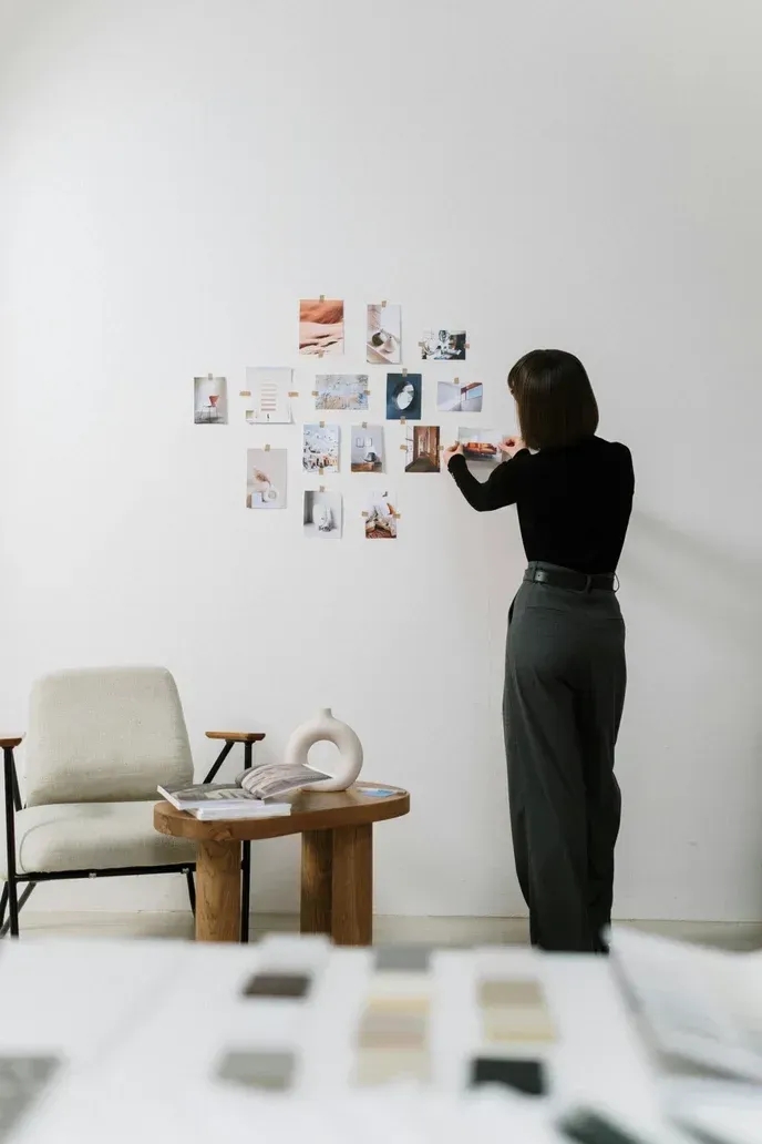 Woman arranging photos on a white wall, near a small table and armchair, with color swatches in foreground.