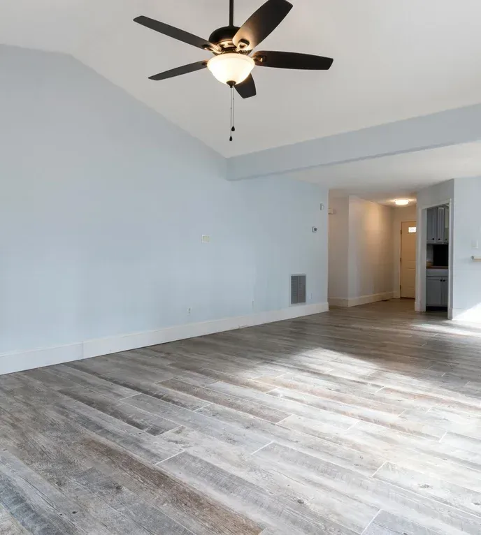Empty room with light blue walls, wood-look floor, ceiling fan, and a doorway leading to a kitchen.